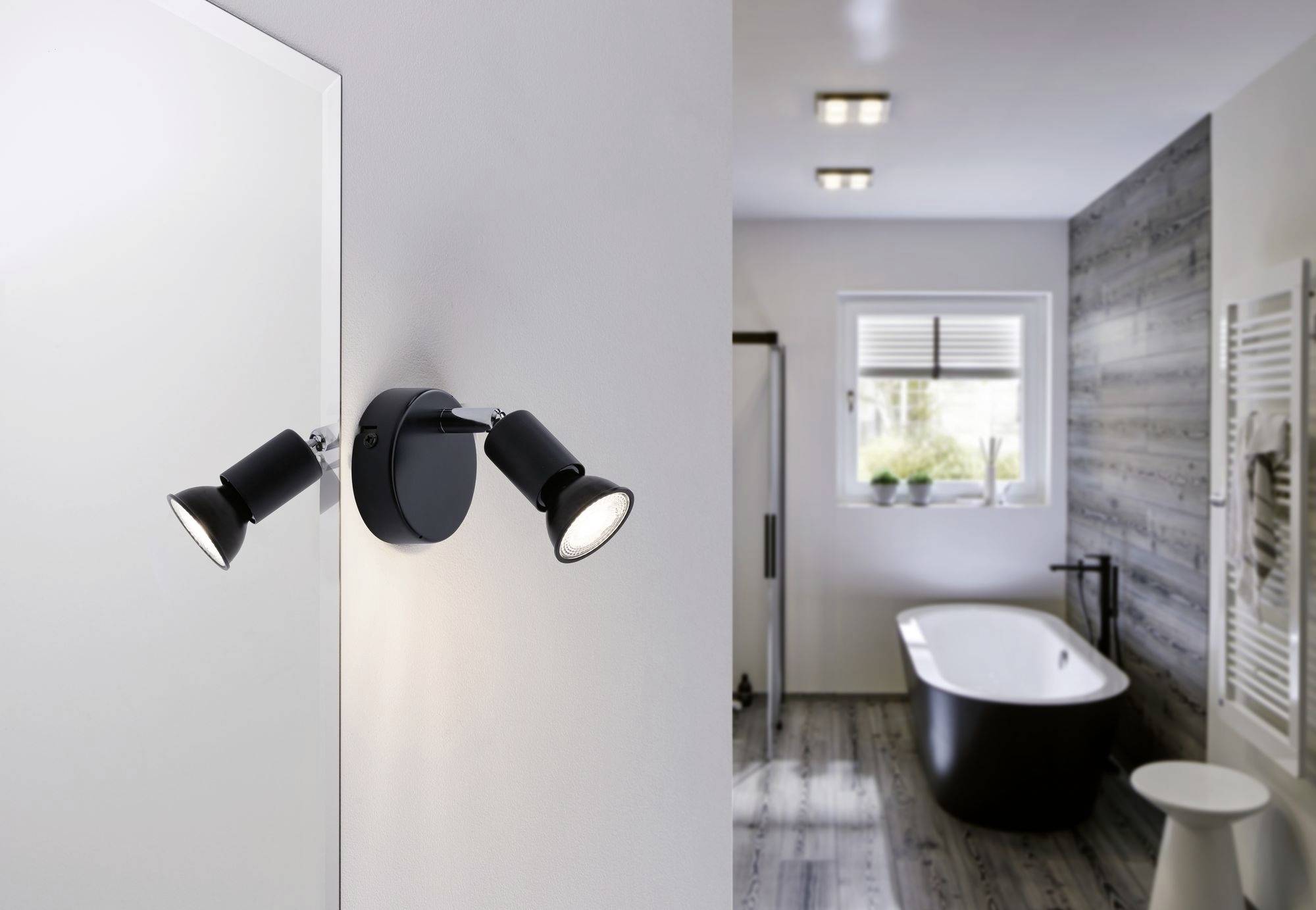 Eine moderne Badezimmerleuchte mit zwei verstellbaren Spots an der Wand. Im Hintergrund ist eine Badewanne und ein Fenster zu sehen.