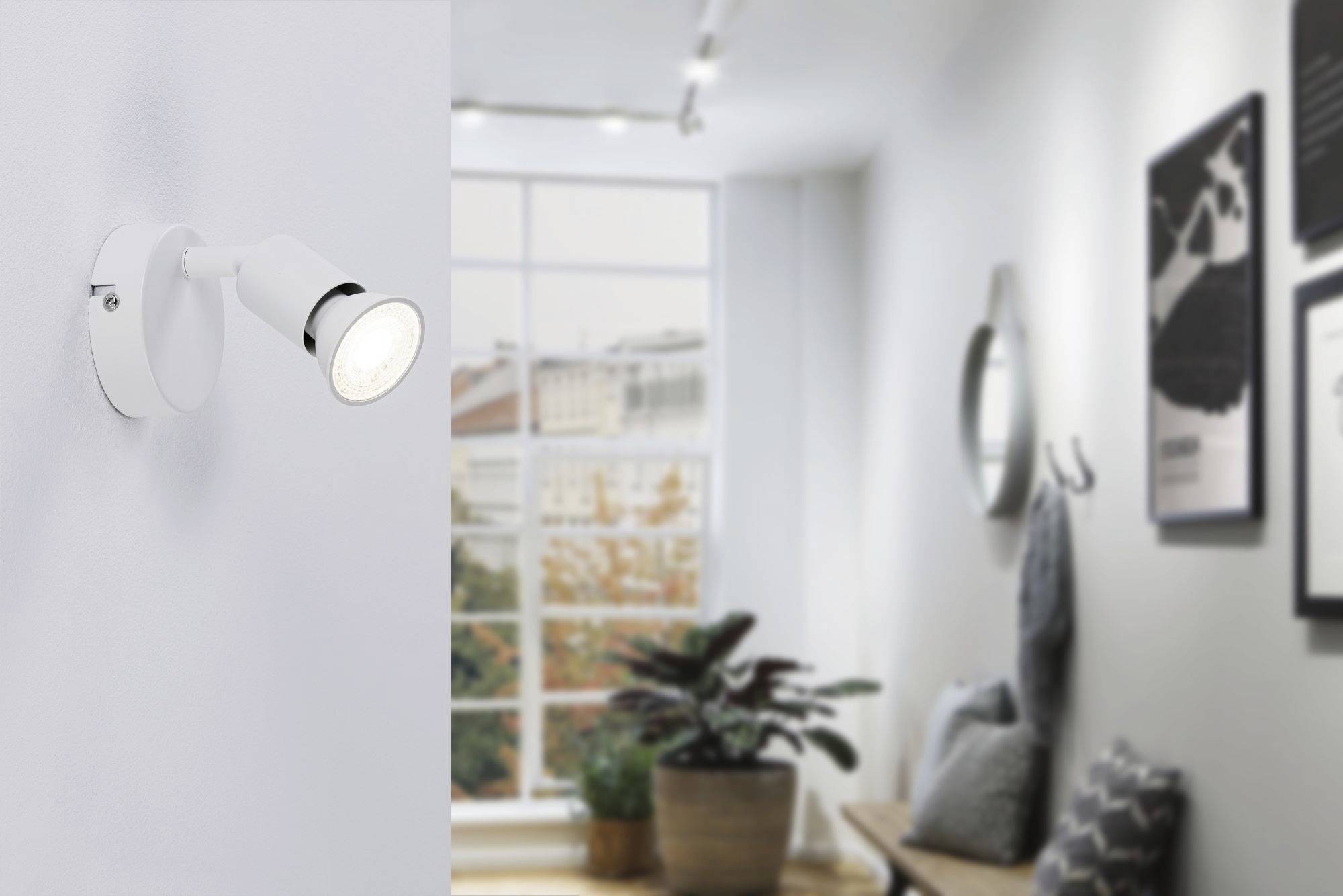 Ein heller, moderner Raum mit einer Wandlampe links. Im Hintergrund sind ein großes Fenster, Pflanzen und gerahmte Bilder an der Wand.