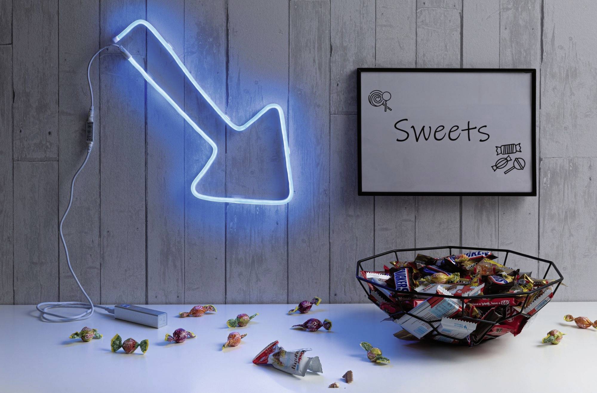 Ein Tisch mit einer Schale voller Süßigkeiten. An der Wand hängt ein leuchtender blauer Pfeil und ein Schild mit der Aufschrift 'Sweets'.