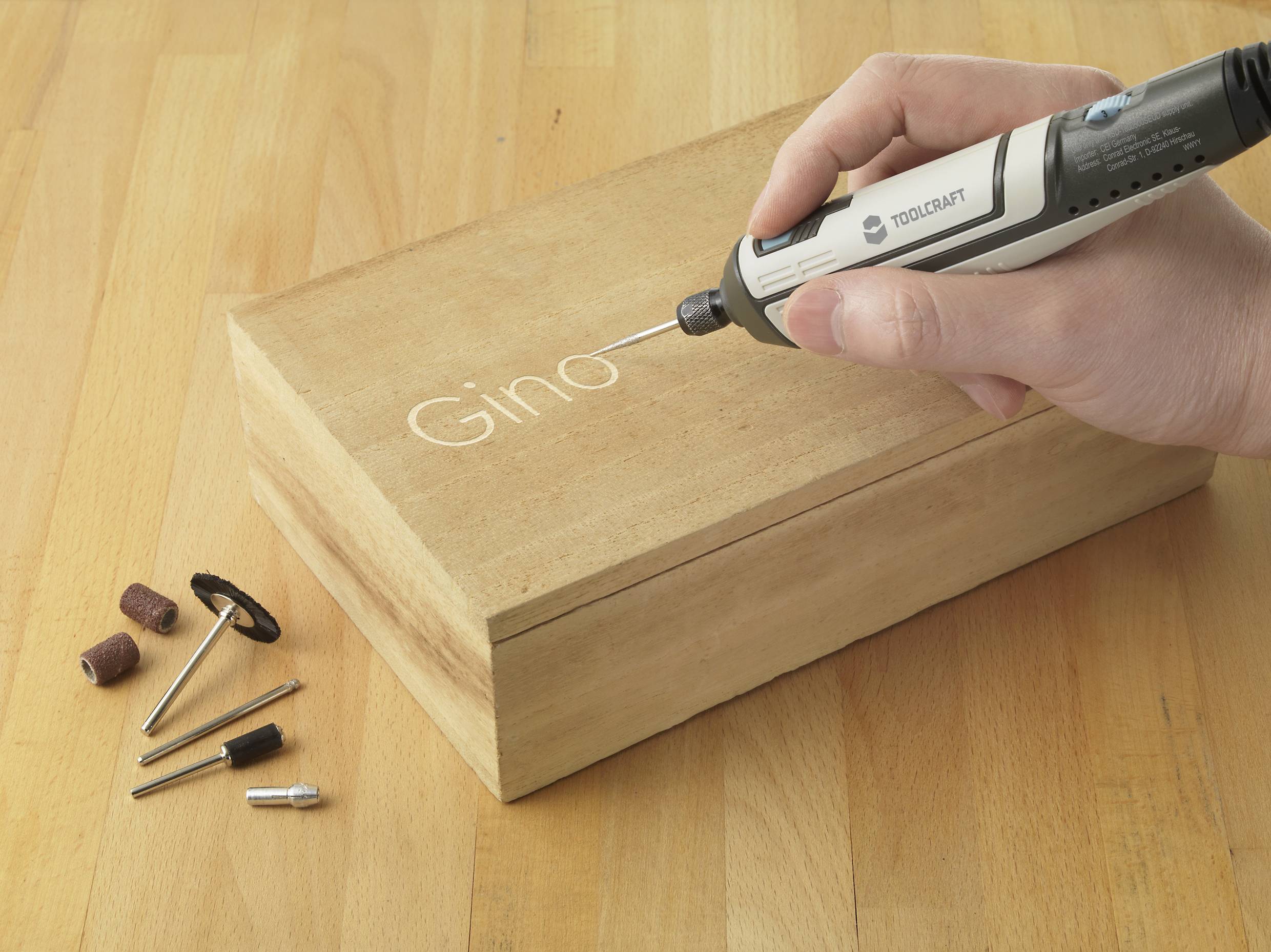 Eine Hand graviert mit einem elektrischen Werkzeug den Namen 'Gino' in ein Holzstück. Daneben liegen unterschiedliche Aufsätze.