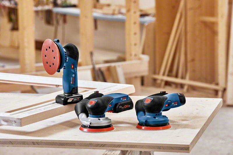 Drei elektrische Exzenterschleifer auf einer Werkbank in einer Holzwerkstatt. Einer steht aufrecht, die anderen liegen flach.