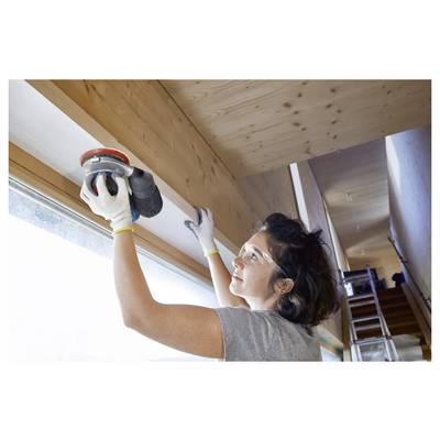 Eine Frau mit Schutzbrille schleift einen Holzbalken in Innenräumen und konzentriert sich dabei sehr konzentriert. Eine Leiter und ein großes Fenster sind im Hintergrund sichtbar und sorgen für natürliches Licht.