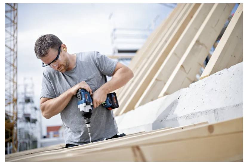 Ein Mann mit Sicherheitsbrille bedient einen Akkuschrauber an einer Holzkonstruktion auf einer Baustelle, umgeben von Gerüsten, unter einem bewölkten Himmel.