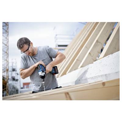 Ein Mann mit Sicherheitsbrille bedient einen Akkuschrauber an einer Holzkonstruktion auf einer Baustelle, umgeben von Gerüsten, unter einem bewölkten Himmel.