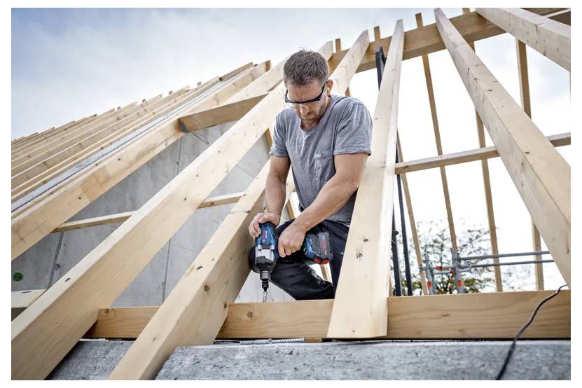 Ein Mann mit Sicherheitsbrille benutzt eine Bohrmaschine auf einem Holzdachrahmen im Bau, mit einem bewölkten Himmel im Hintergrund.