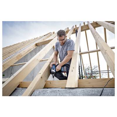 Ein Mann mit Sicherheitsbrille benutzt eine Bohrmaschine auf einem Holzdachrahmen im Bau, mit einem bewölkten Himmel im Hintergrund.
