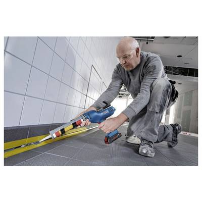 Eine Person kniet auf einem gefliesten Boden und verwendet eine blaue elektrische Fugenpistole, um die Kante zwischen dem Boden und den Wandfliesen zu versiegeln.