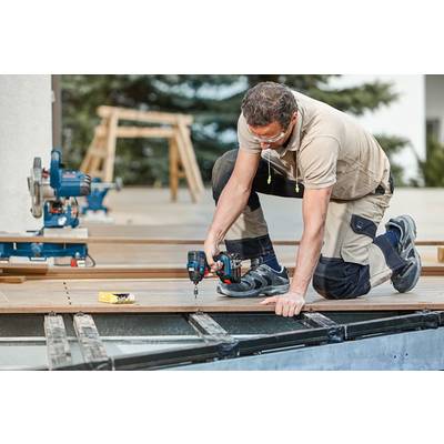 Ein Mann kniet und benutzt eine Bohrmaschine, um Holzdielen zu befestigen. Im Hintergrund sind Werkzeuge und eine Leiter sichtbar.