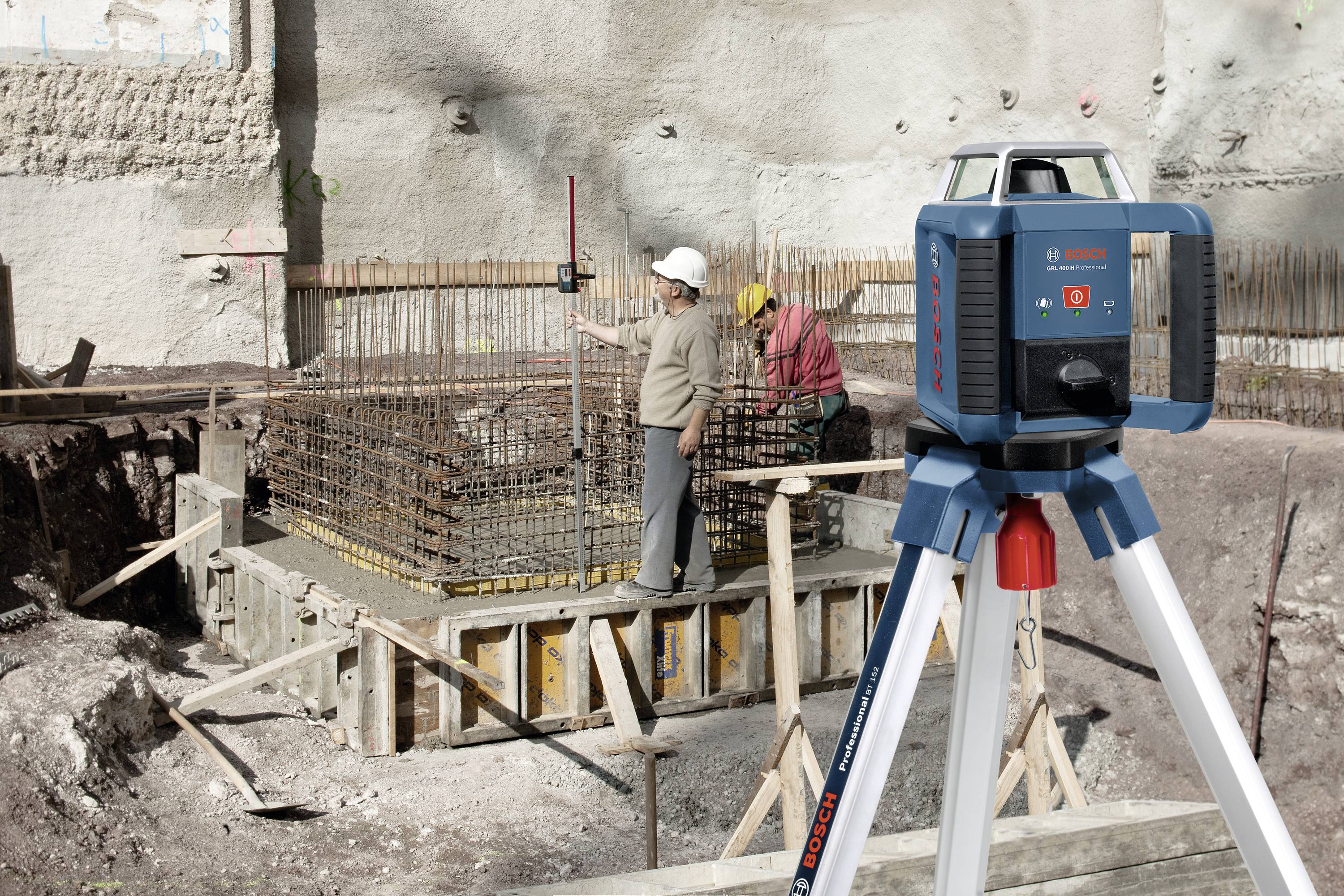 Bauarbeiter verwenden Nivelliergeräte auf einer Baustelle; sie messen die Höhe einer Betonfundamentbefestigung. Im Vordergrund steht ein blau-graues Lasergerät auf einem weißen Stativ.