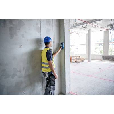 Ein Bauarbeiter mit Schutzhelm und Warnweste misst mit einem Gerät die Wand eines Rohbaus. Umgebung zeigt unvollendeten Bau.