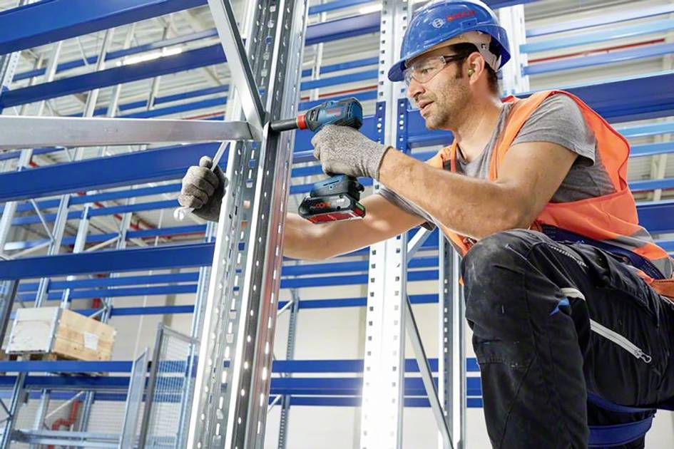 Ein Arbeiter in Sicherheitsausrüstung befestigt Metallträger mit einem Bohrschrauber in einem Lager.