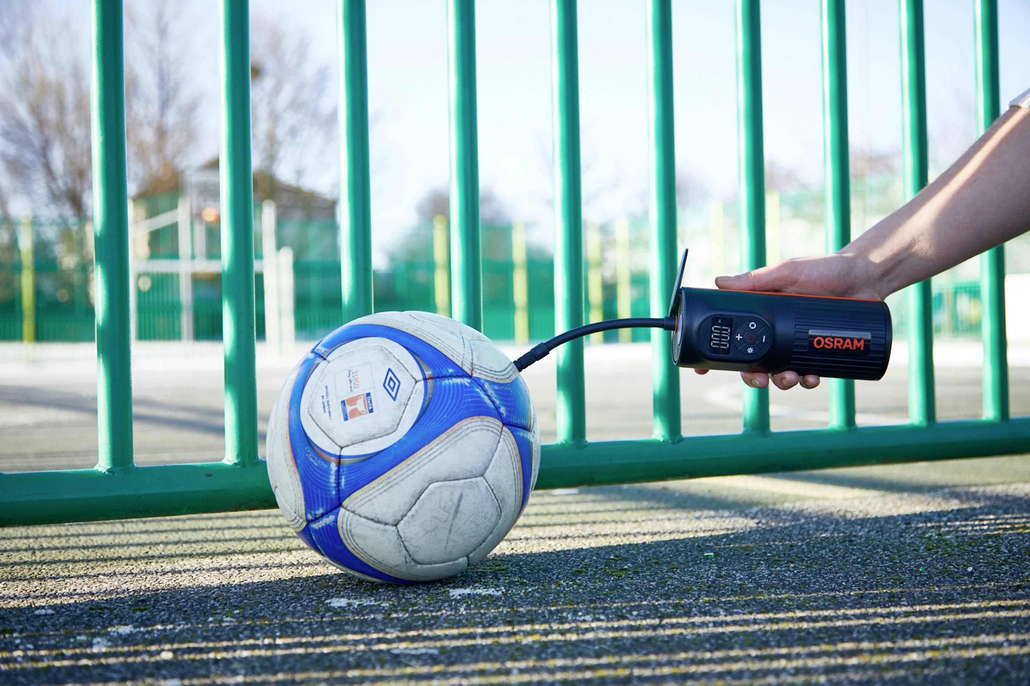 Eine Hand hält eine Luftpumpe von Osram, die mit einem Ventil an einem Fußball befestigt ist. Der Ball liegt auf einem Sportplatz mit grünem Zaun.
