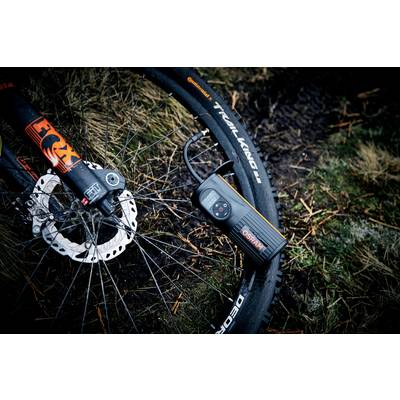 Fahrradreifen mit einer schwarzen elektrischen Luftpumpe auf schlammigem Boden im Freien aufgepumpt. Links sichtbar ist die Fahrradgabel.