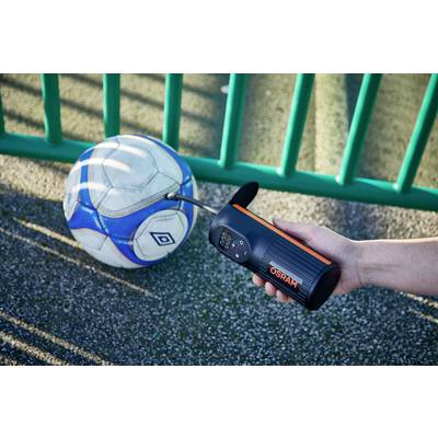 Eine Person pumpt einen Fußball mit einem tragbaren Gerät auf, während der Ball auf einer gepflasterten Oberfläche neben einem grünen Zaun liegt.