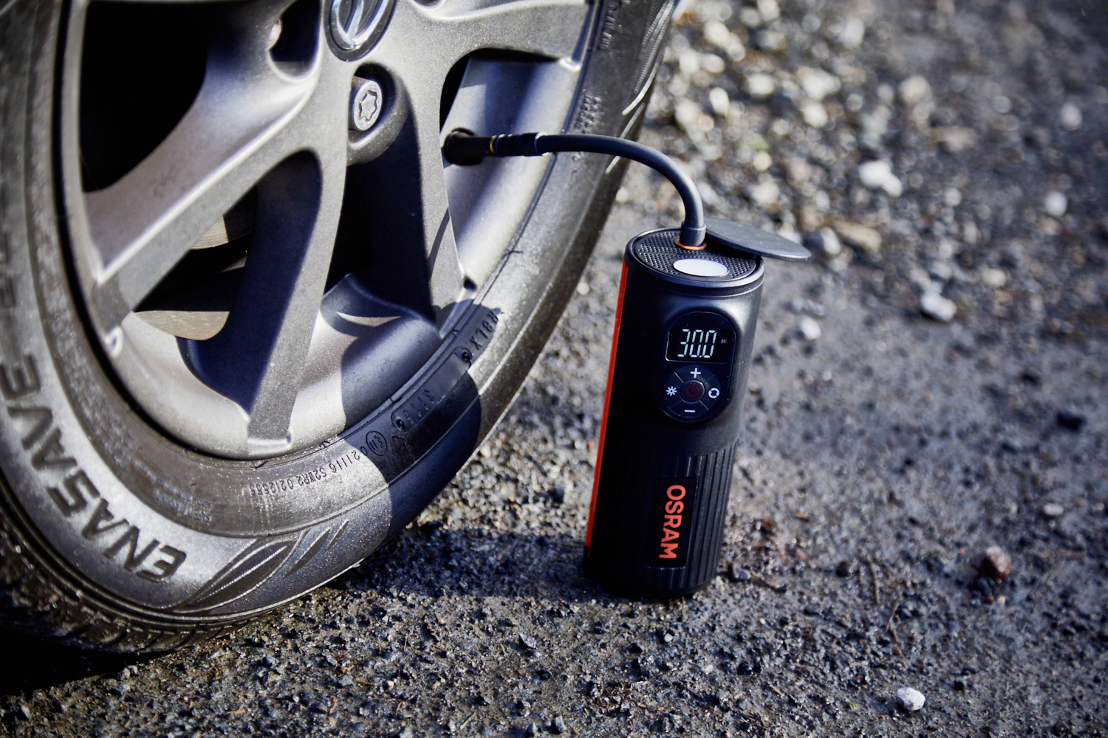 'Kleiner digitaler Luftkompressor mit Anzeige von 3,00 bar an einen Autoreifen angeschlossen, auf einer asphaltierten Oberfläche.'