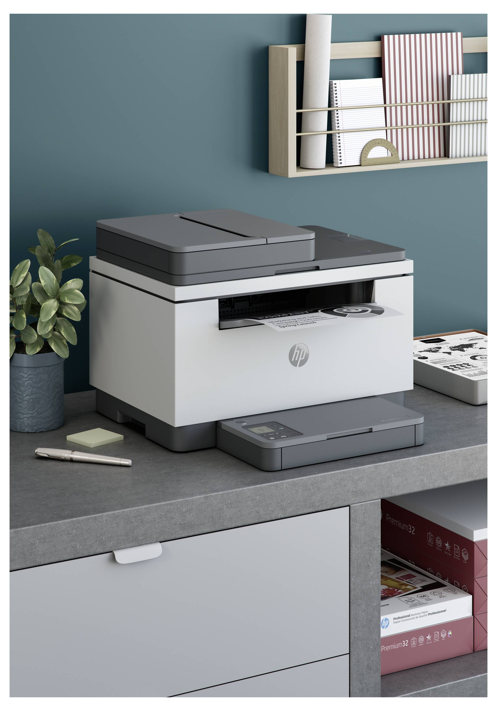Ein kompakter HP-Multifunktionsdrucker sitzt auf einem Schreibtisch in einem Heimbüro, mit einer Pflanze, Stiften, Papieren und Wandregalen in der Nähe.