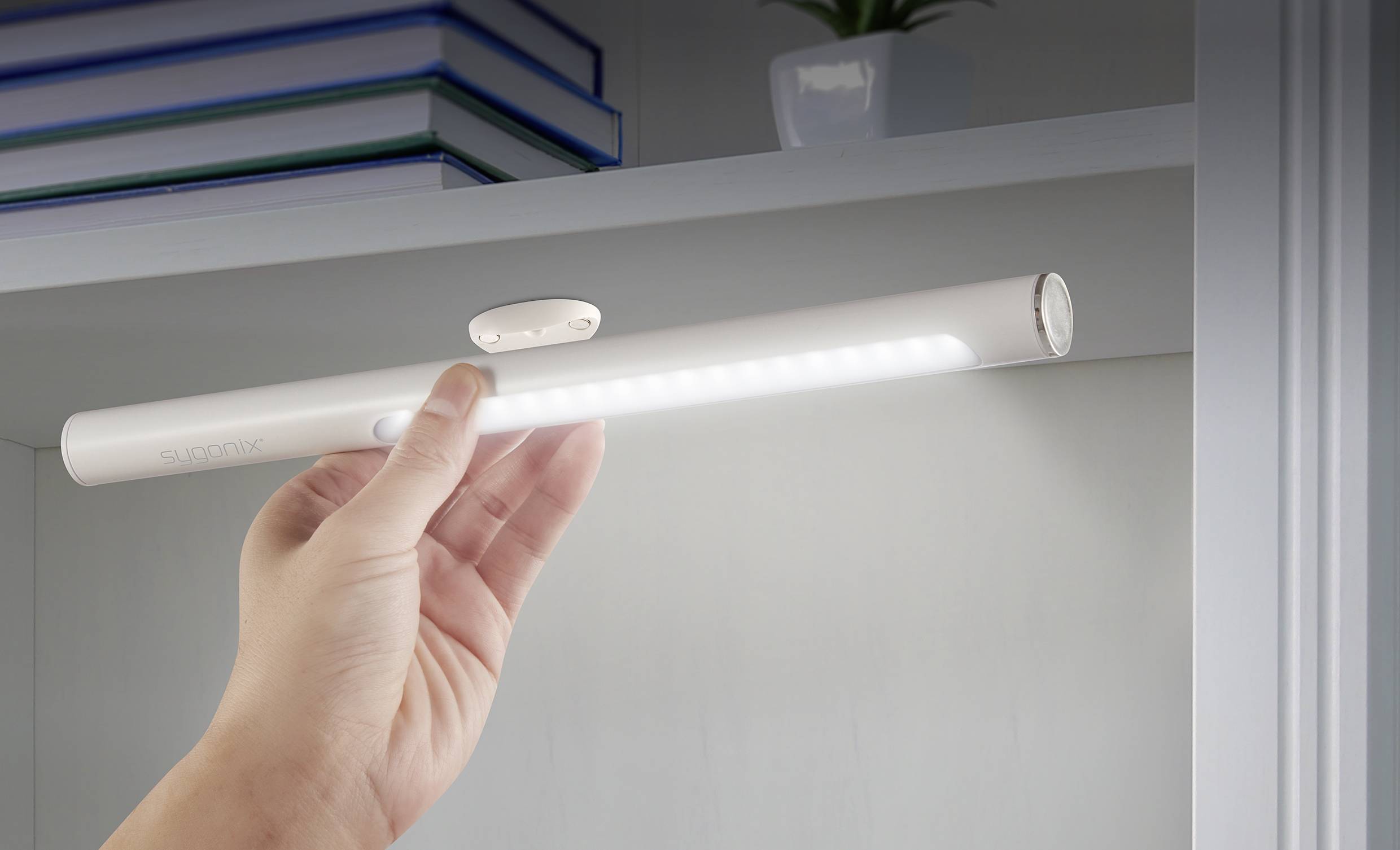 Eine Hand hält ein schlankes, batteriebetriebenes LED-Nachtlicht unter einem Regal. Im Hintergrund sind Bücher und eine Pflanze zu sehen.