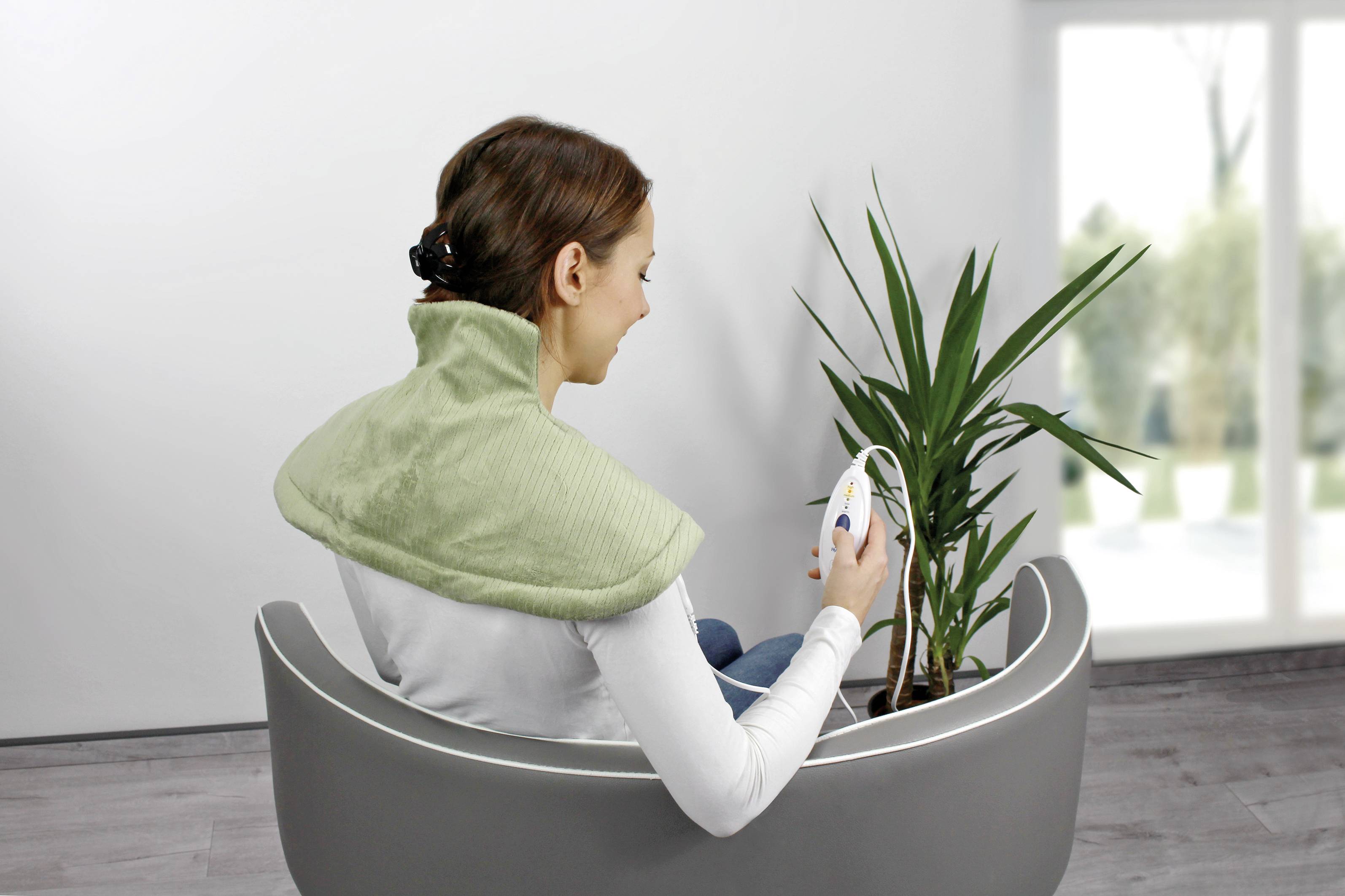 Eine Frau sitzt in einem modernen Stuhl mit einem grünen Wärmekissen um den Nacken und hält ein Gerät, neben ihr steht eine Pflanze.