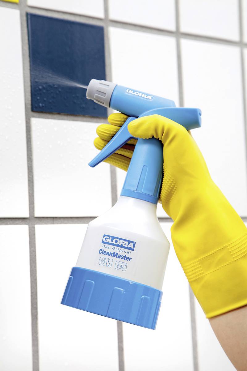 Eine Hand in einem gelben Gummihandschuh hält einen blauen und weißen Sprüher und besprüht eine geflieste Wand mit Reinigungsmittel.