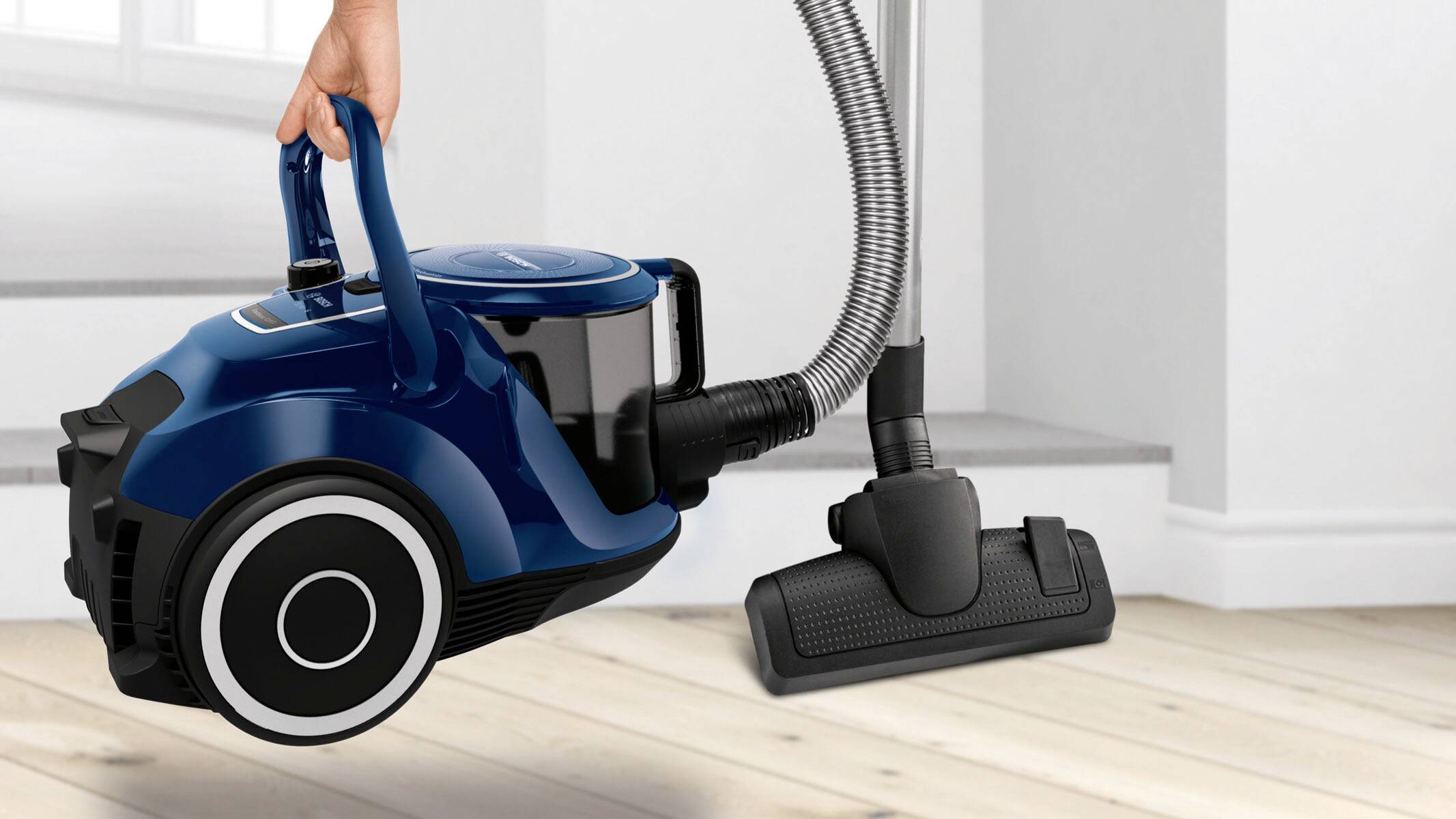 Person hebt einen blauen Staubsauger in einem Raum mit hellem Holzboden an. Das Gerät hat ein modernes, kompaktes Design.