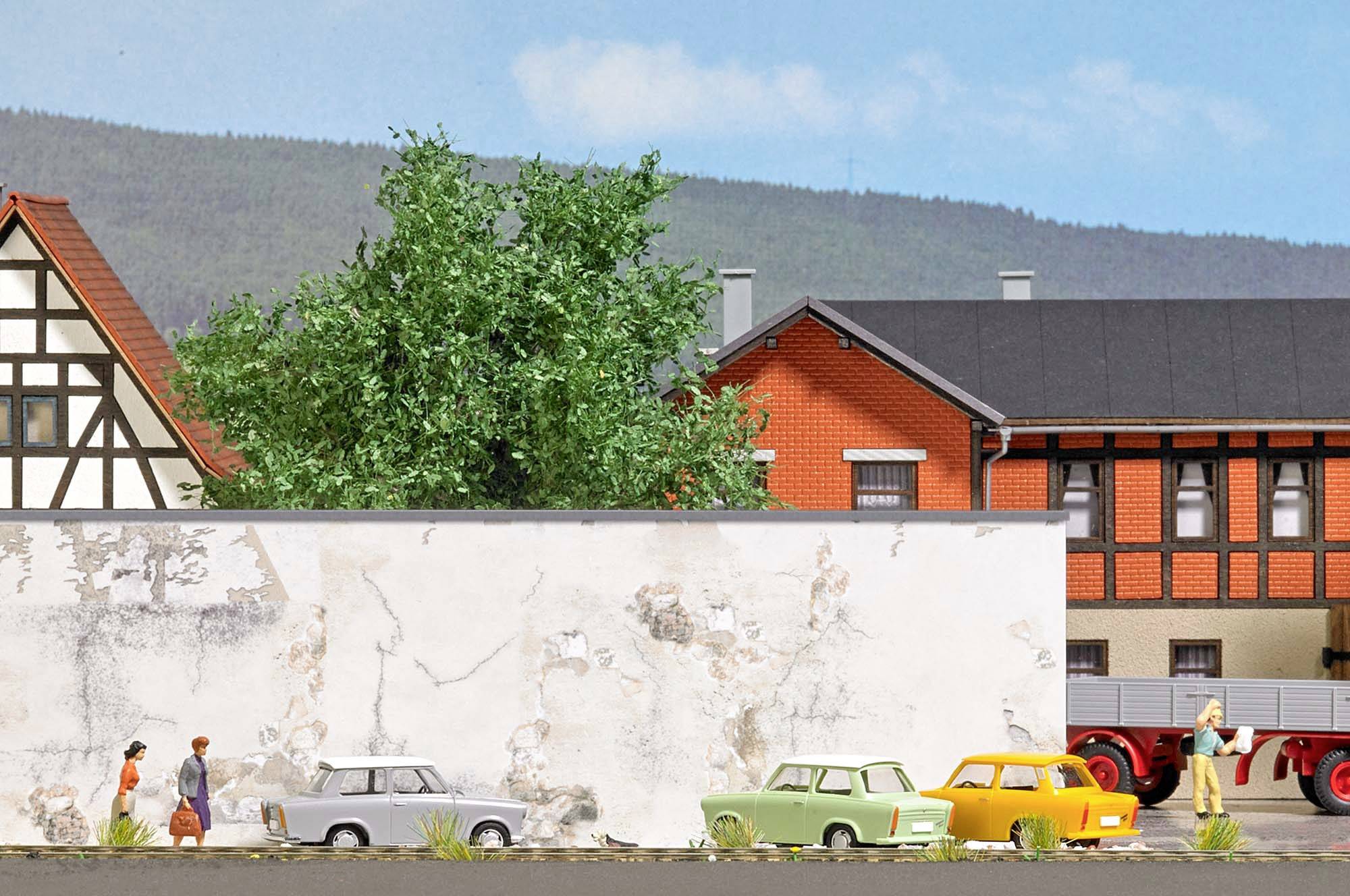 Eine Mauer trennt eine Straße mit geparkten Autos von zwei historischen Gebäuden. Zwei Personen gehen an der Mauer entlang.