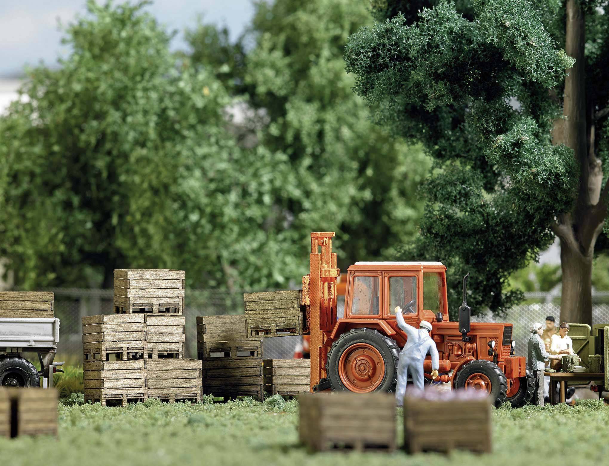 Eine Miniaturszene zeigt einen roten Traktor neben Holzkisten, umgeben von grünen Bäumen und Figuren, die im Hintergrund arbeiten.