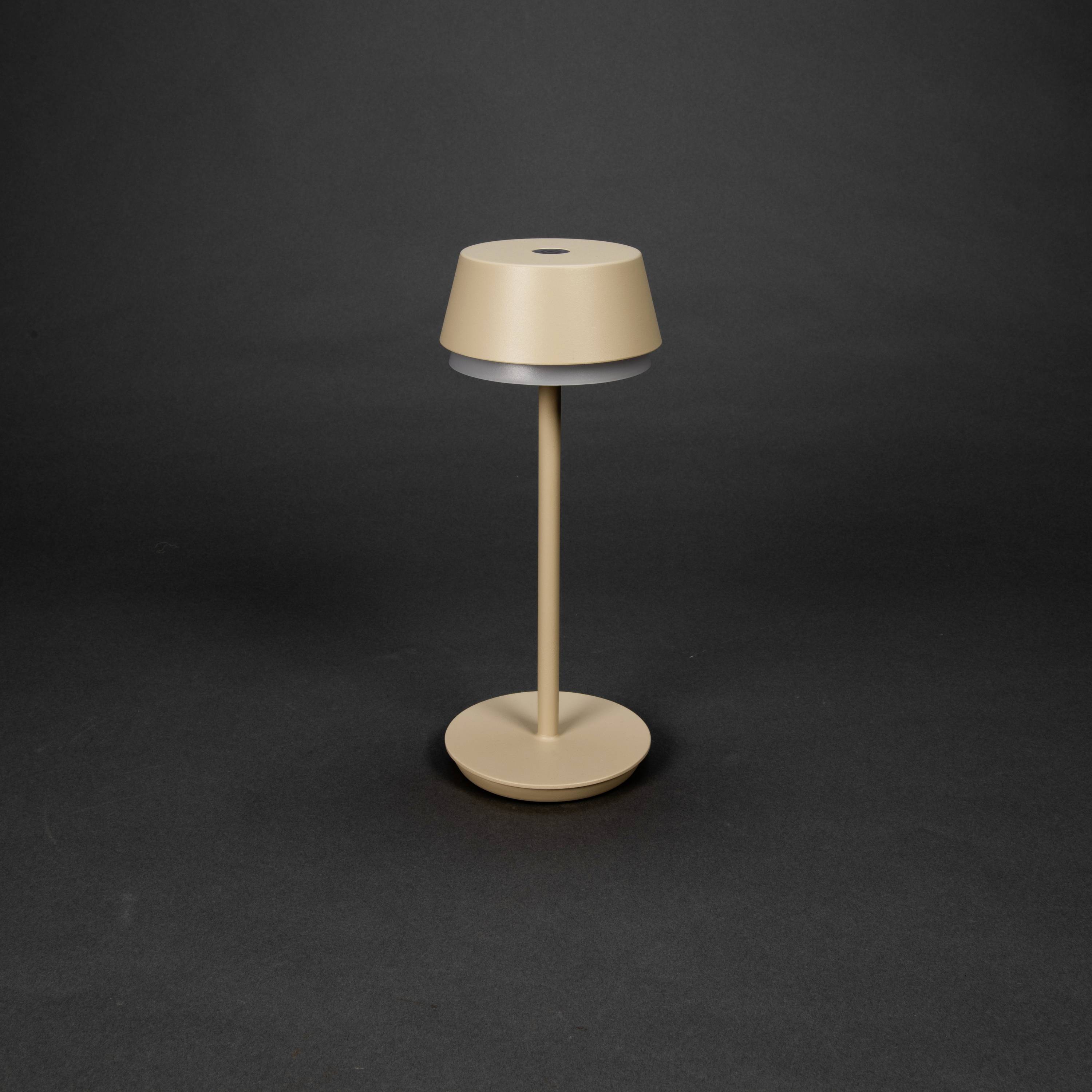 Eine minimalistische, beige Tischlampe auf dunklem Hintergrund. Sie hat einen runden Schirm und einen schlanken Stiel auf einer kreisförmigen Basis.