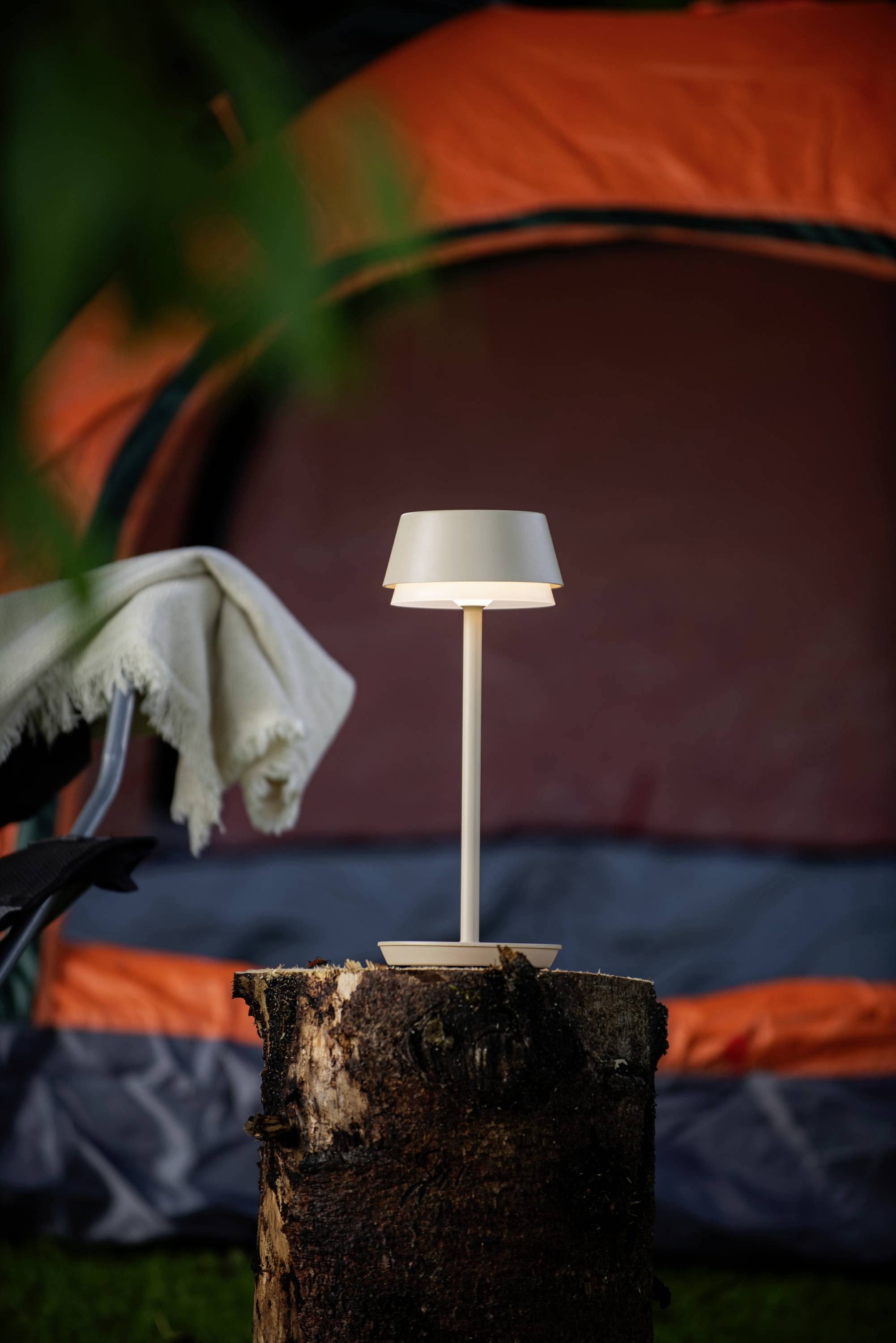 Eine Lampe steht auf einem Baumstumpf vor einem orangefarbenen Zelt. Daneben liegt eine Decke über einem Campingstuhl.