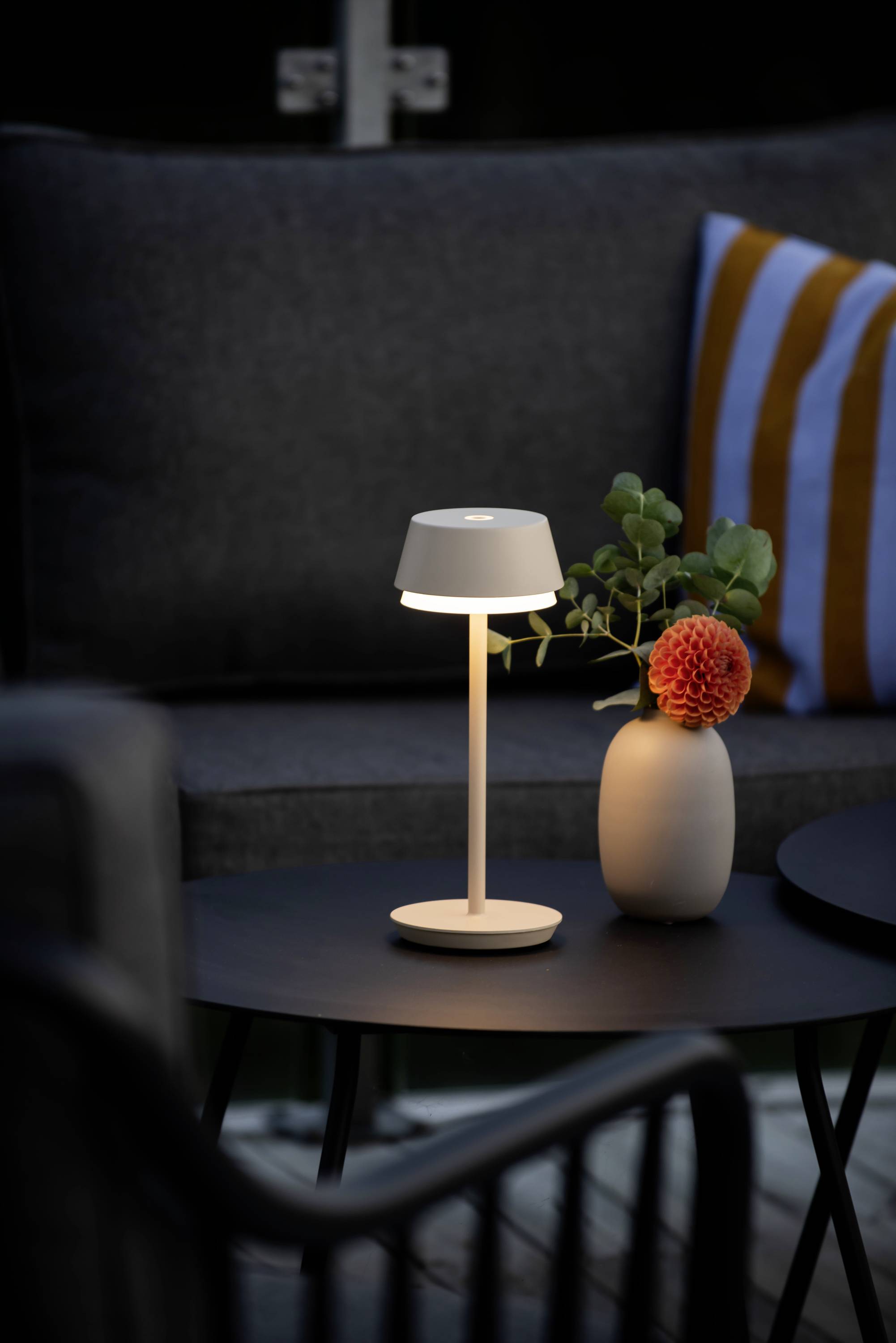 Kleine Tischlampe leuchtet auf einem runden Tisch neben einer Vase mit oranger Blume. Im Hintergrund ist ein graues Sofa.