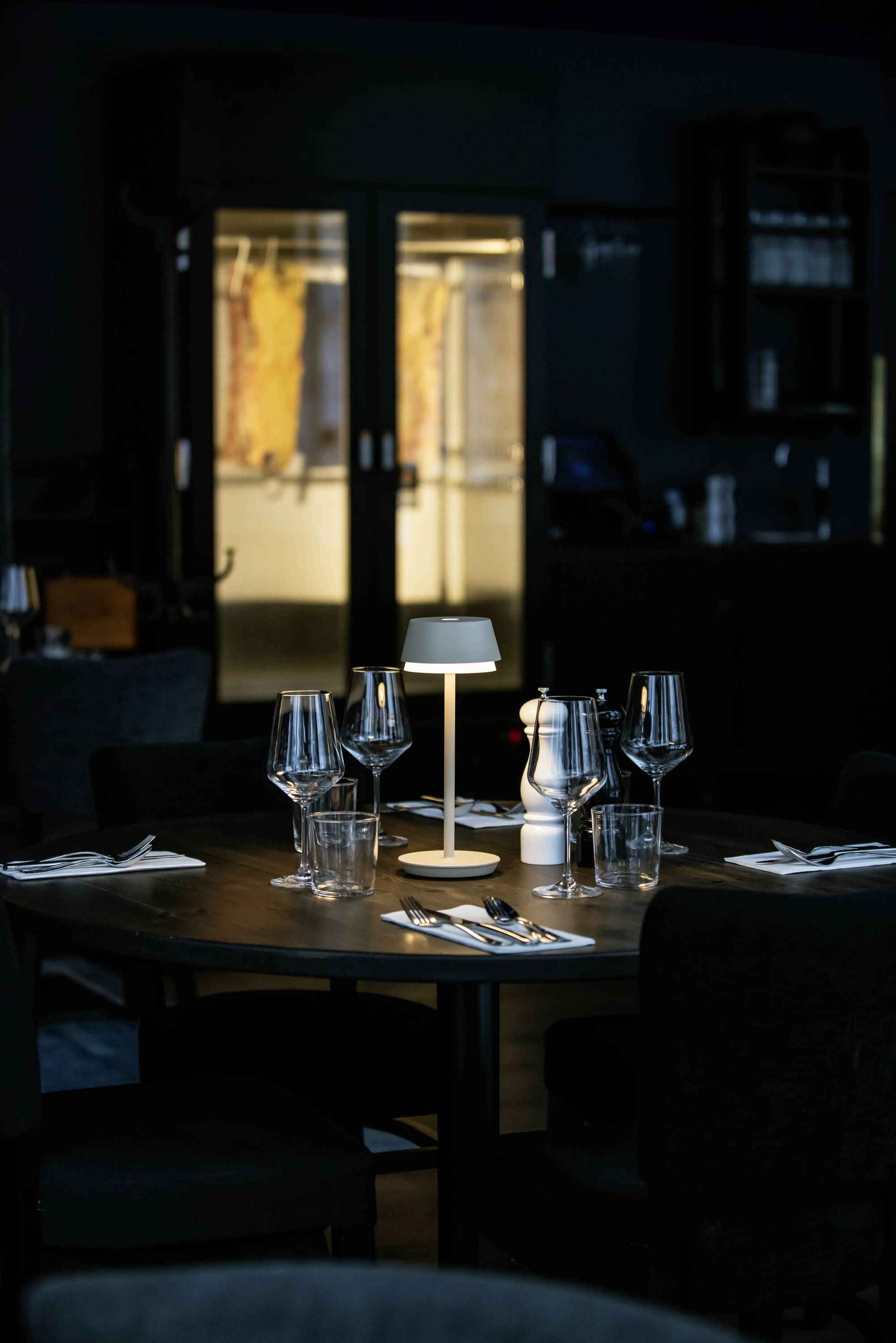 Ein stilvoll gedeckter Holztisch in einem dunklen Restaurant, beleuchtet von einer kleinen Tischlampe. Im Hintergrund ist ein Weinschrank sichtbar.
