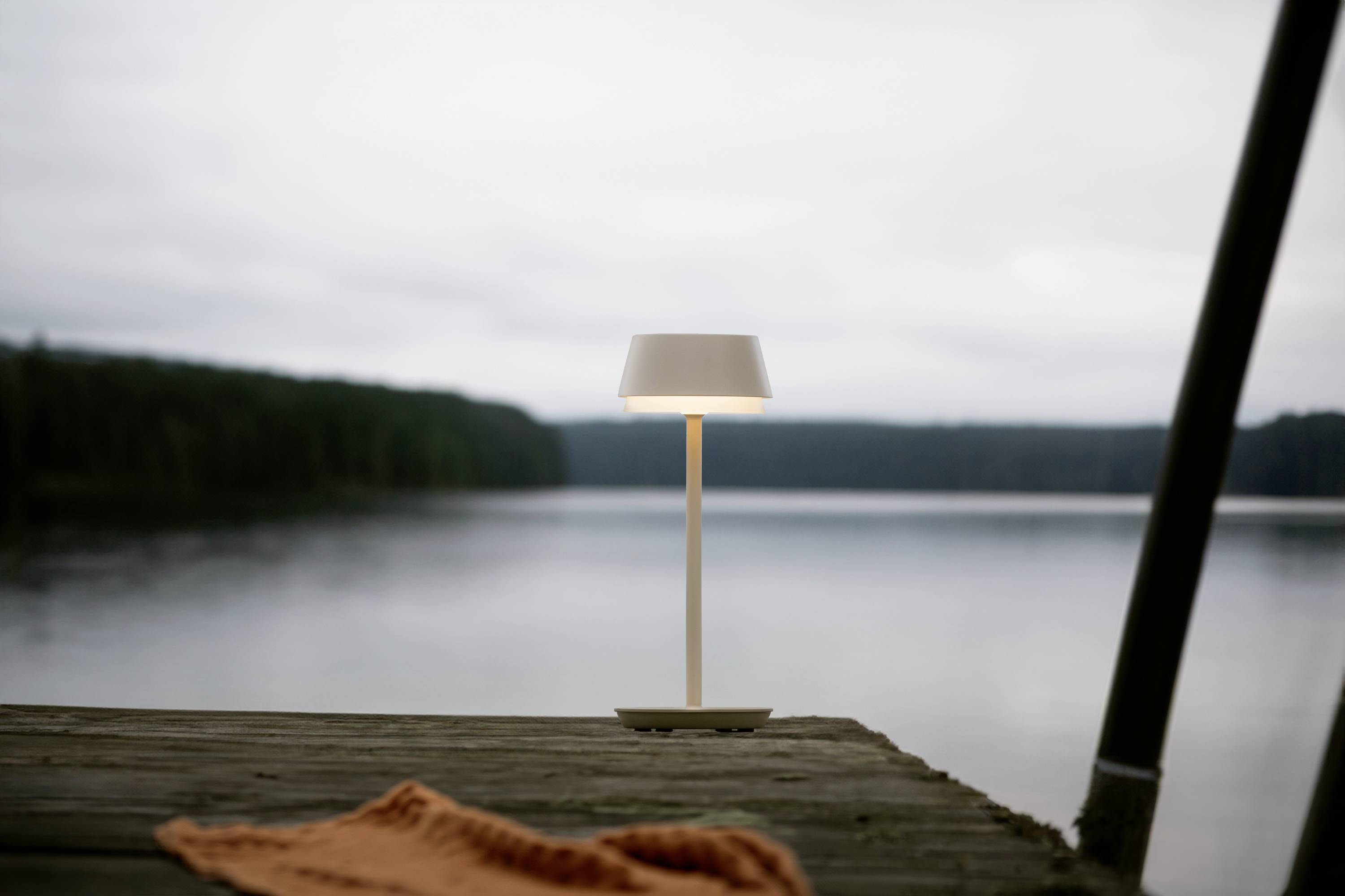 Eine Lampe steht auf einem Holzdock vor einem ruhigen See. Der Himmel ist bewölkt und Wald erstreckt sich am Horizont. Ein orangefarbenes Tuch liegt auf dem Dock.
