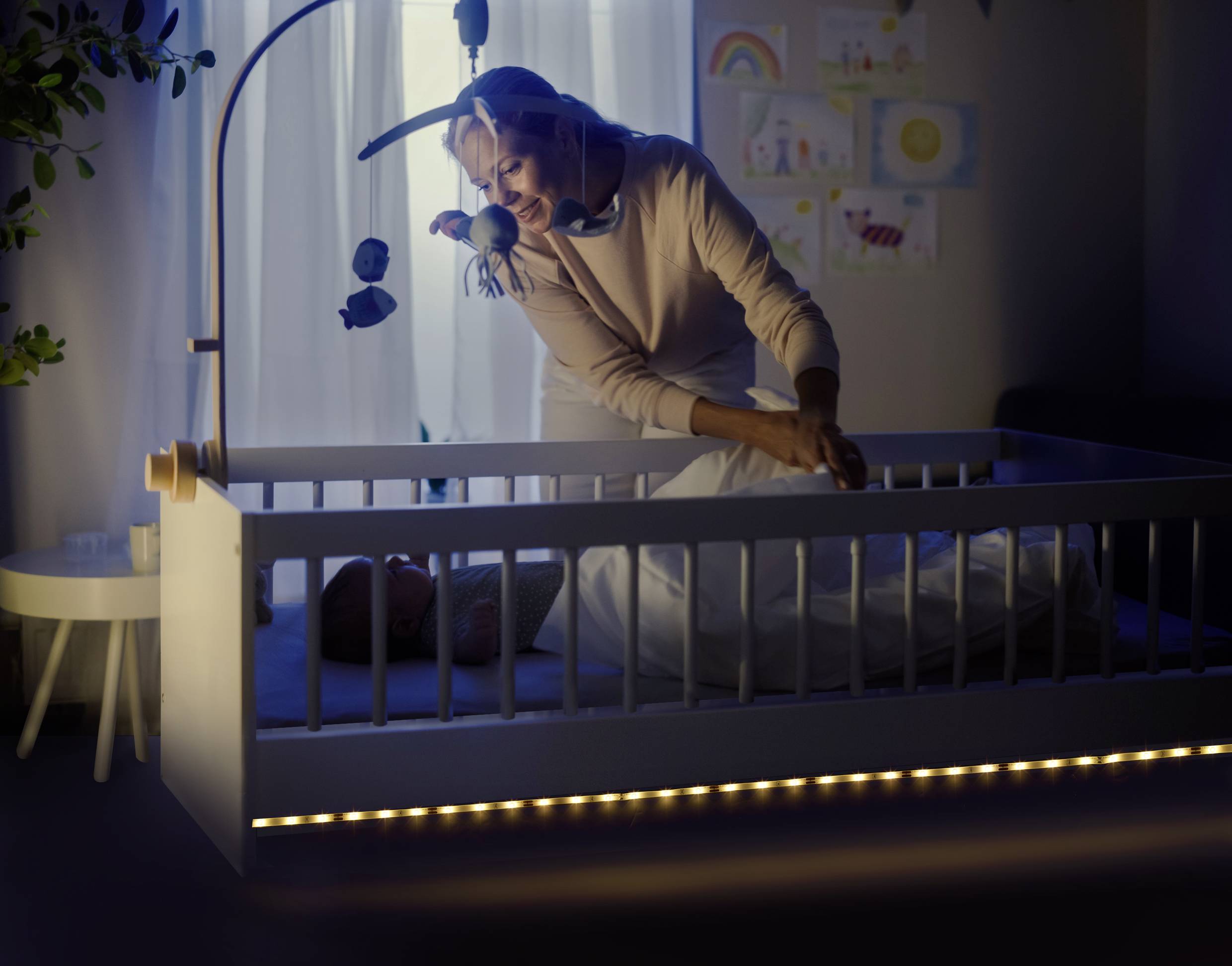 Eine Frau im gedämmten Raum beugt sich über ein beleuchtetes Babybett und deckt ein Baby zu. Im Hintergrund sind Kinderzeichnungen zu sehen.
