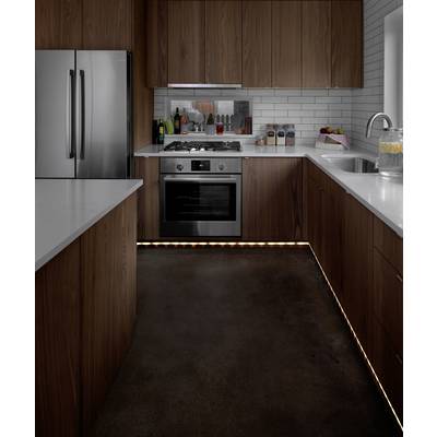 Moderne Küche mit Holzschränken, weißer Arbeitsplatte, Edelstahlgeräten und Beleuchtung am Boden. Links steht ein Kühlschrank neben einem Herd.