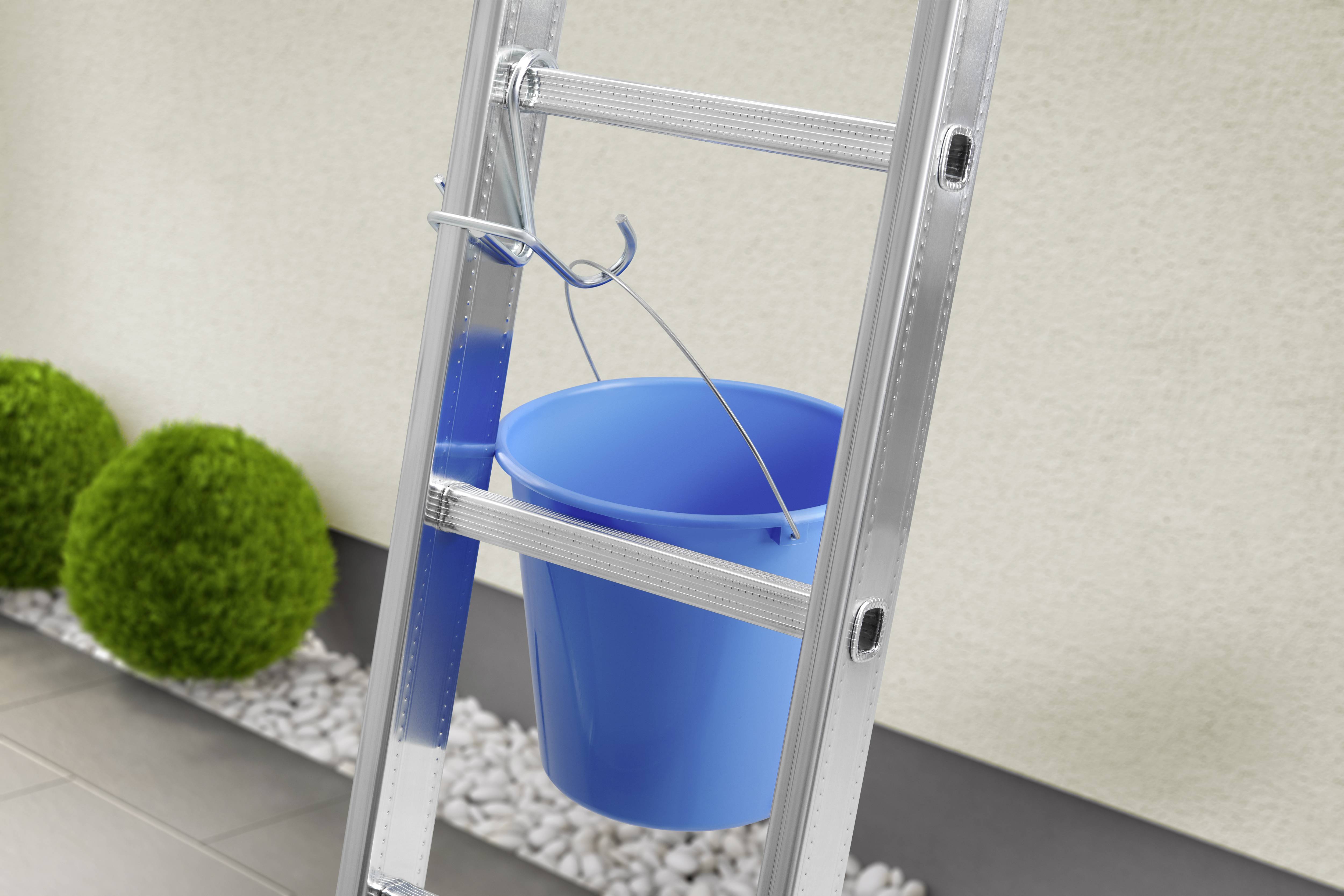 Eine blaue Plastikeimer hängt an einer Leiter vor einer Wand, umgeben von Kieselsteinen und grünen Büschen.