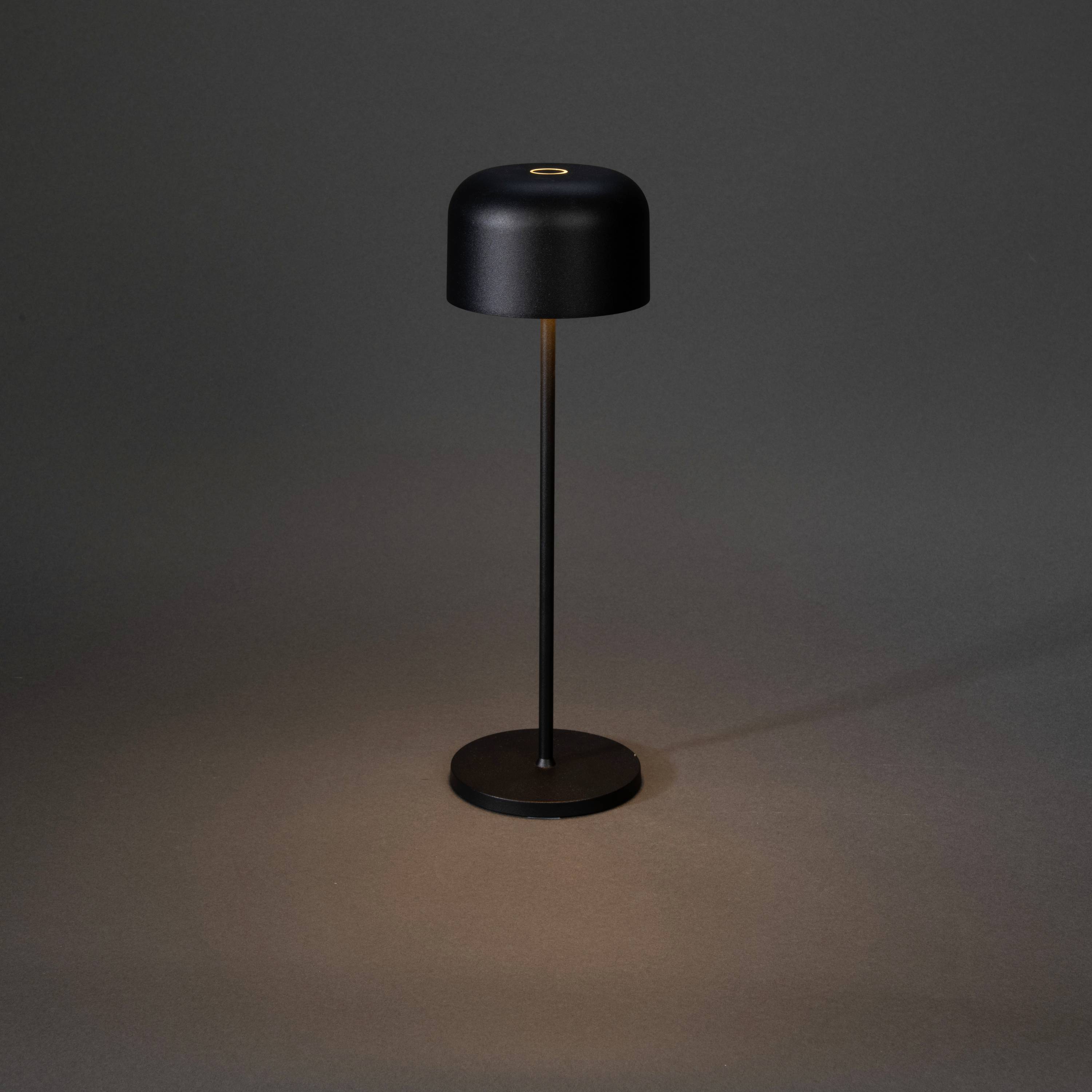 Moderne schwarze Tischlampe mit rundem Schirm und Standfuß, leuchtet schwach auf dunklem Hintergrund.