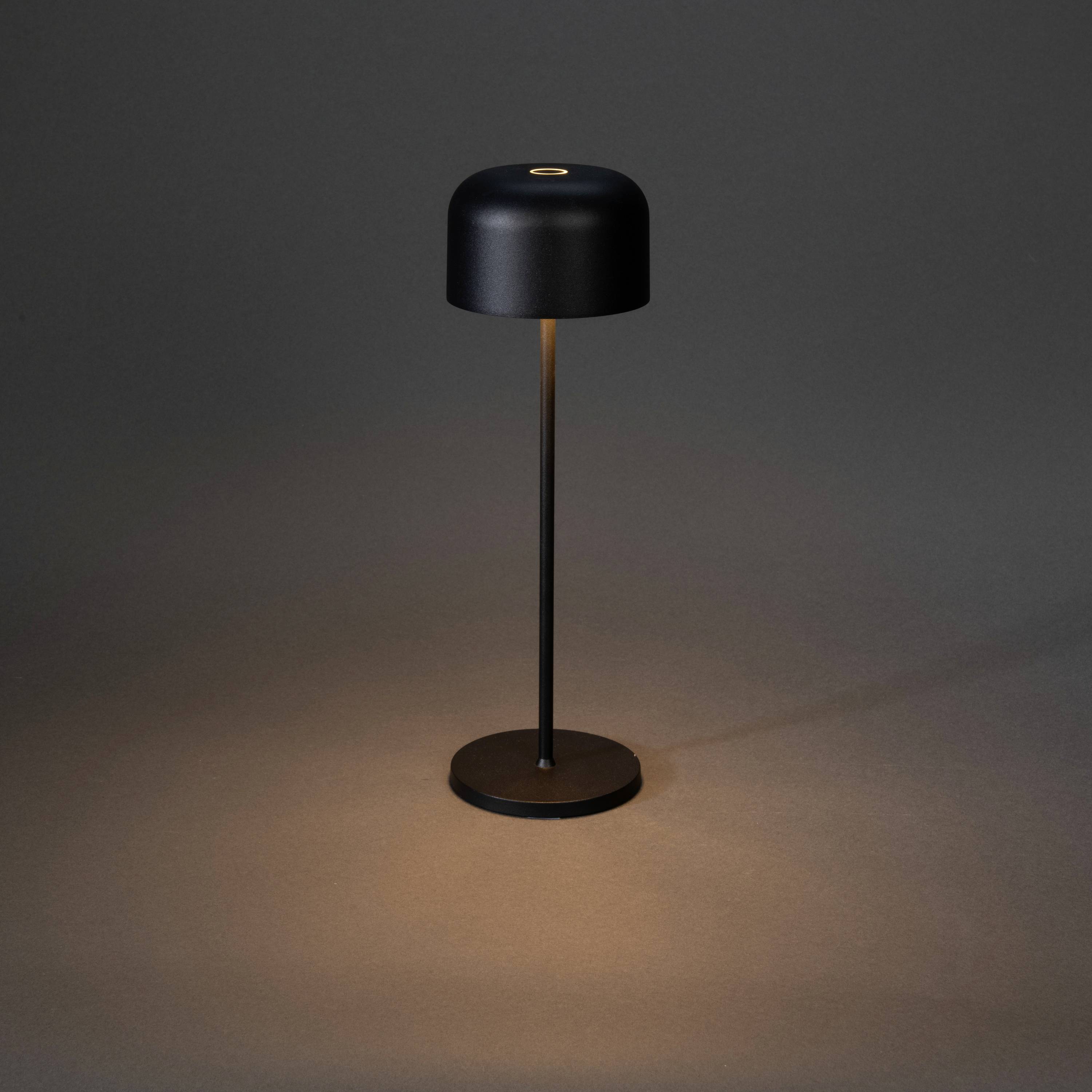 Eine schwarze Tischlampe mit rundem Schirm steht auf einem glatten, dunklen Untergrund und wirft warmes Licht darunter.