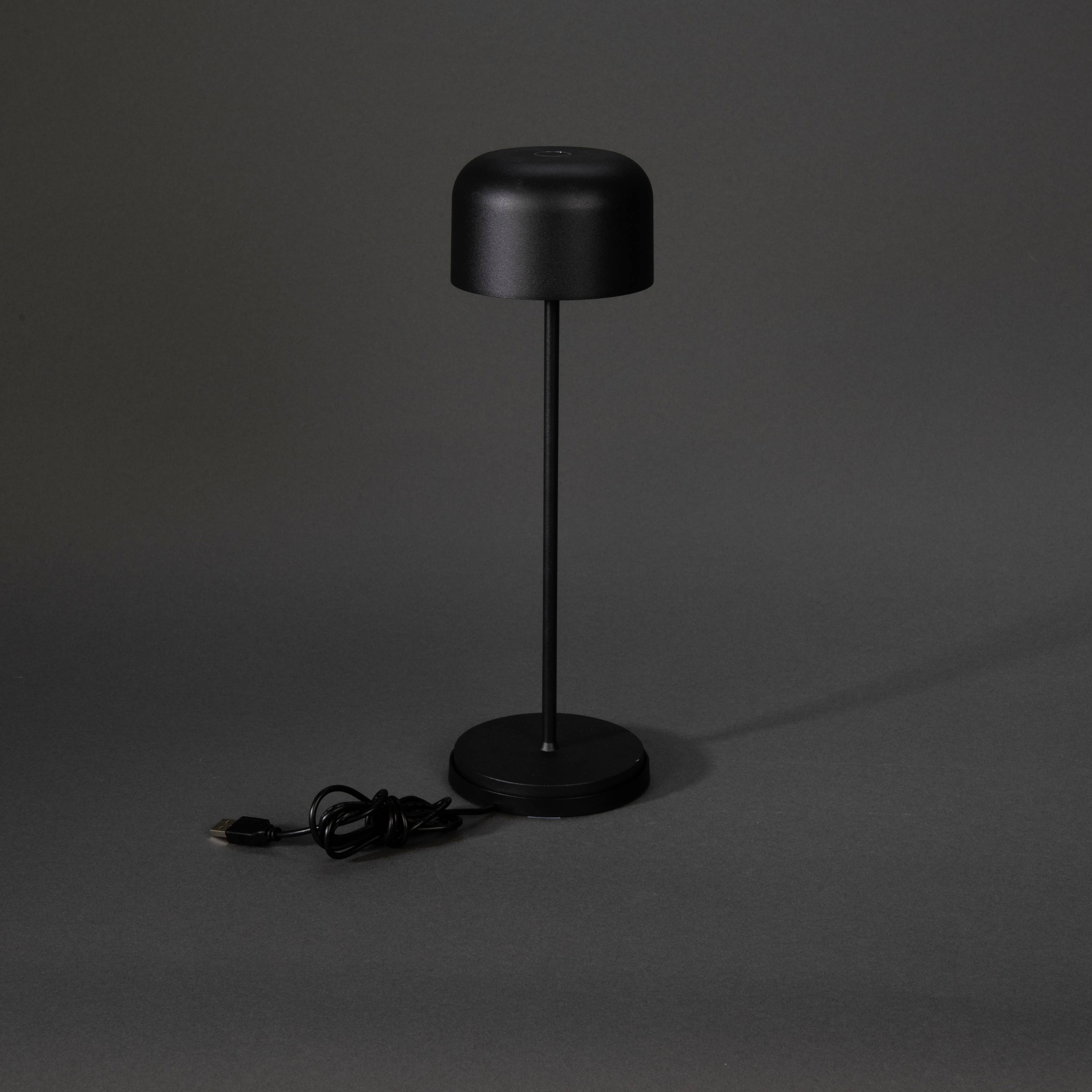 Schwarze Tischlampe mit schlichtem, modernem Design. Der Lampenschirm ist zylindrisch, der Sockel rund. Kabel liegt rechts daneben.