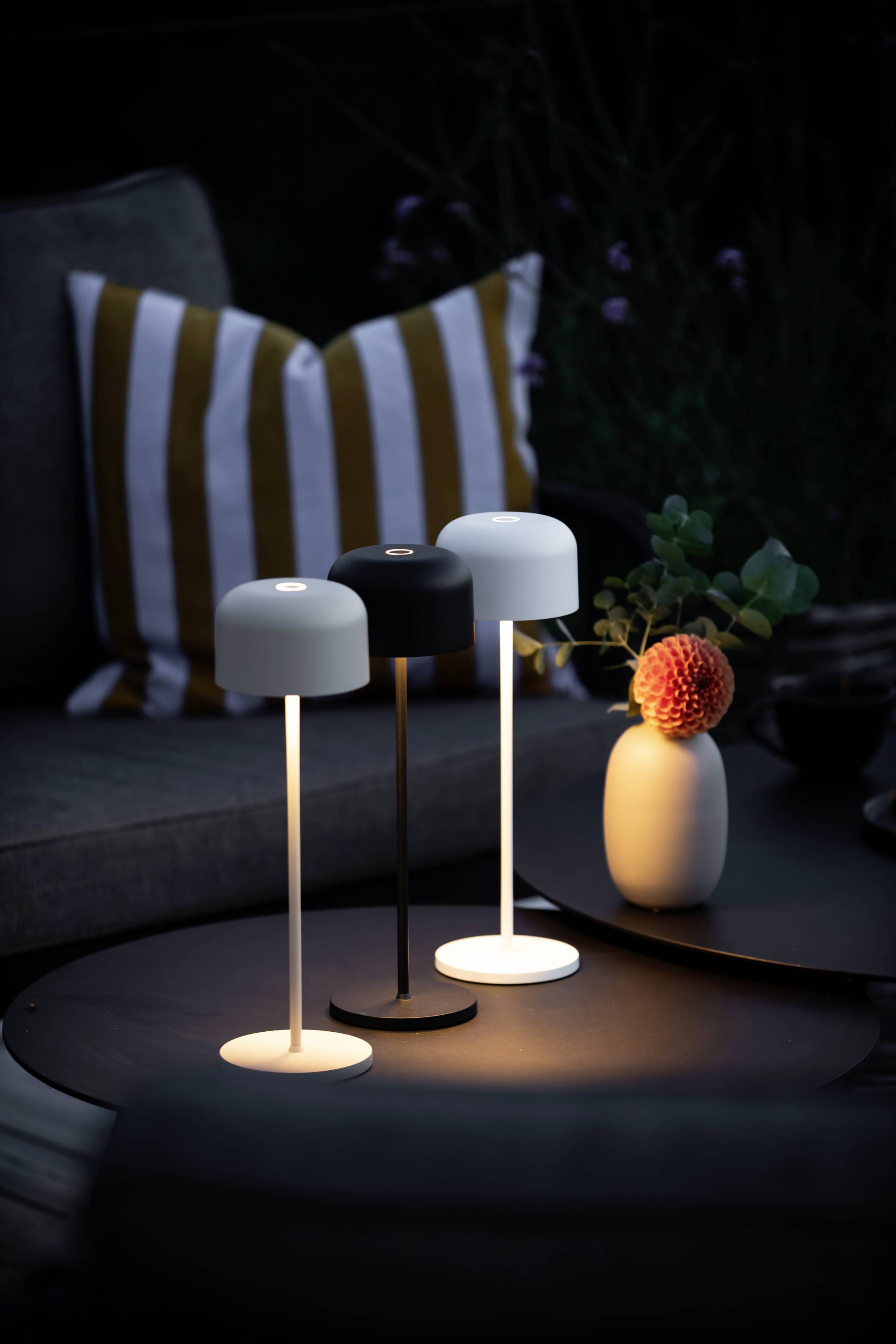 Drei moderne Tischlampen in Weiß und Schwarz leuchten auf einem runden Tisch; dahinter ein gestreiftes Kissen und eine Vase mit Blume.