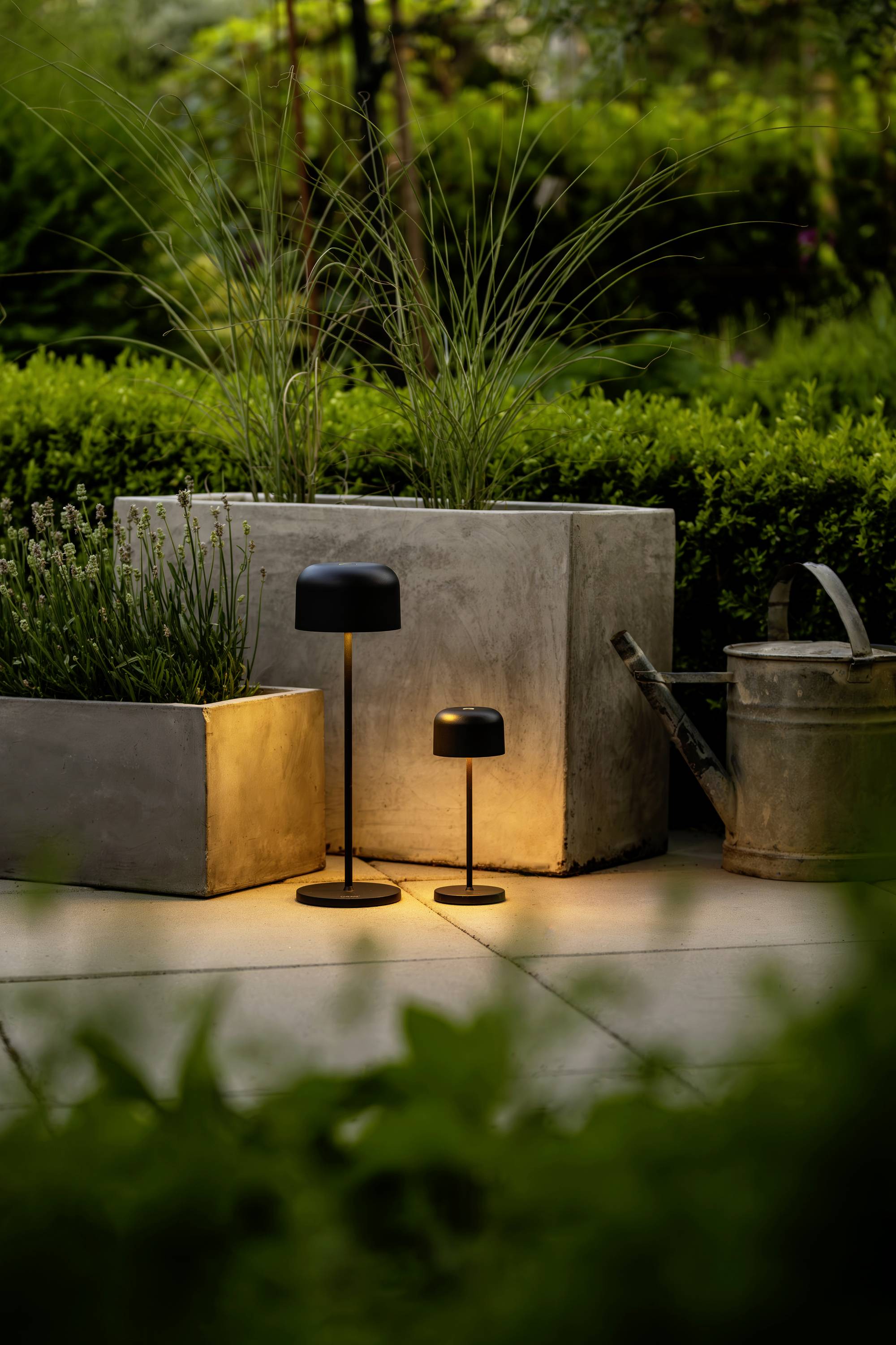 Zwei moderne, schwarze Außenlampen leuchten neben rechteckigen Pflanzkübeln auf einer Terrasse, umgeben von grünen Pflanzen.
