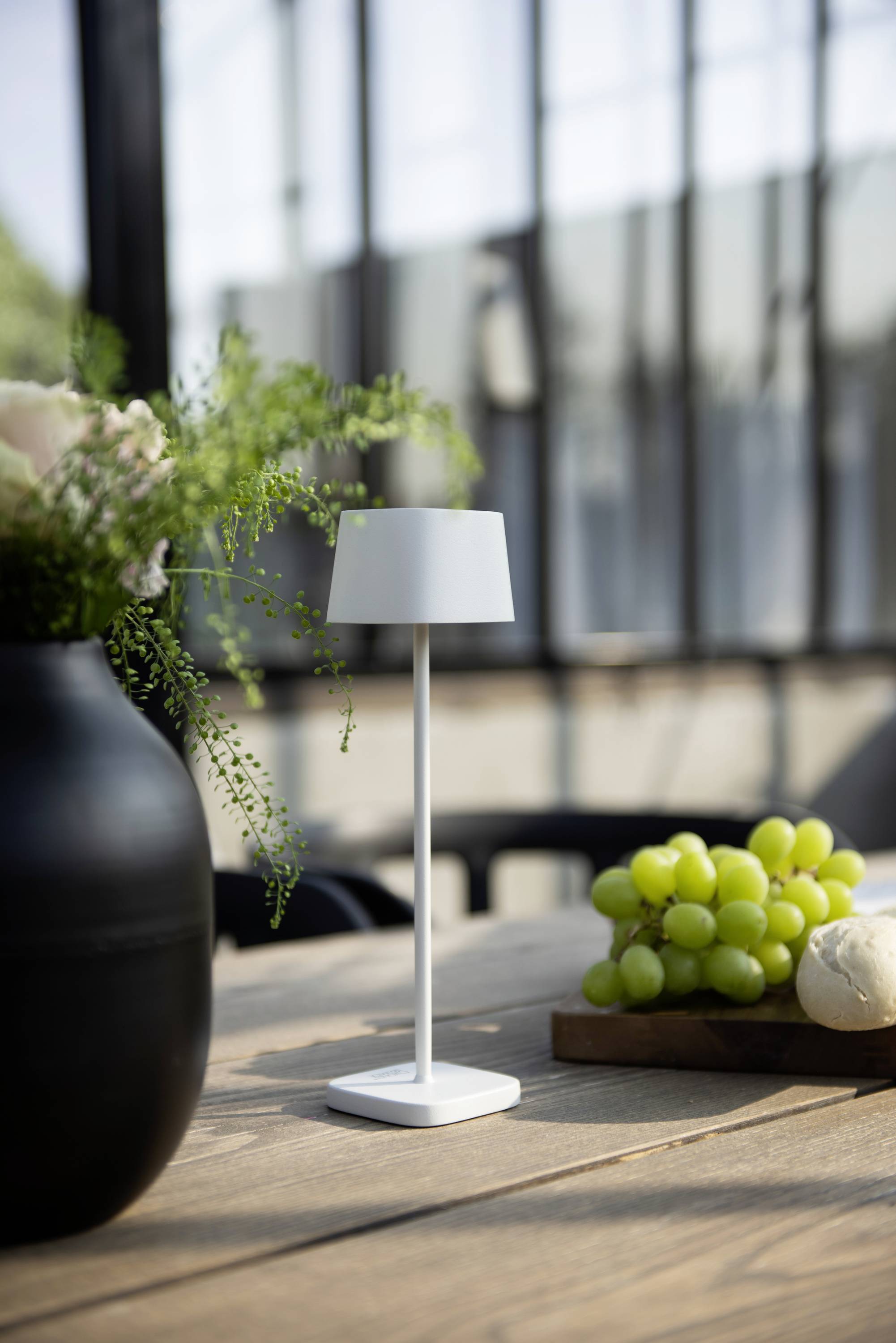 Eine moderne Tischlampe steht neben einem Blumenarrangement in einer Vase und einem Tablett mit grünen Trauben auf einem Holztisch.