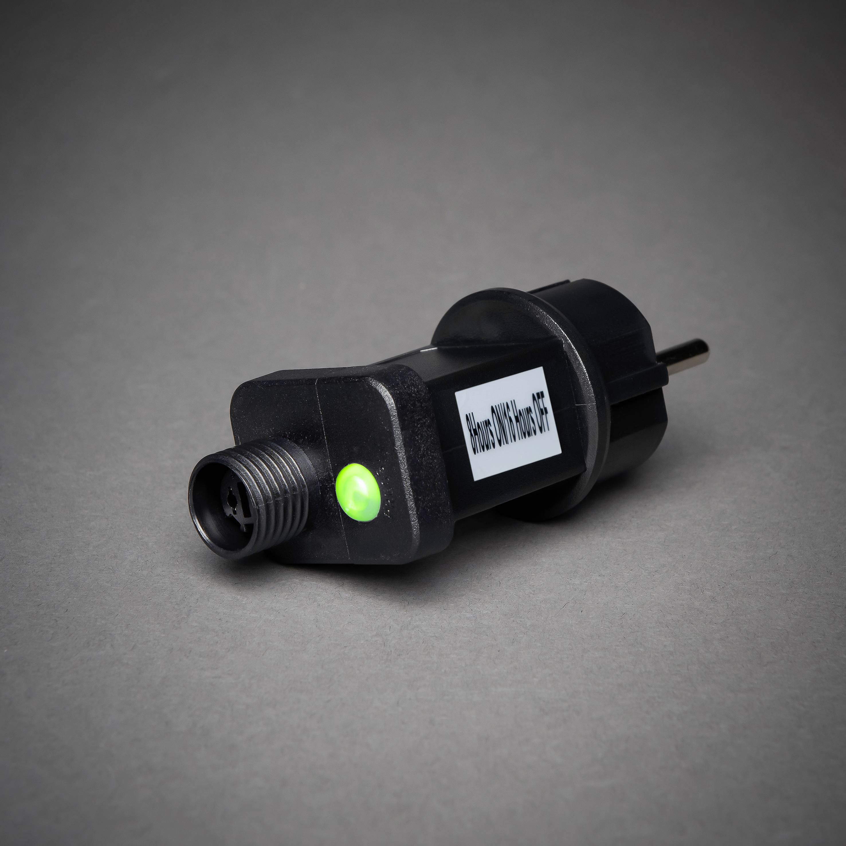 Ein schwarzer Stromstecker mit einem grün leuchtenden Kontrolllicht und einem weißen Etikett auf grauem Hintergrund.