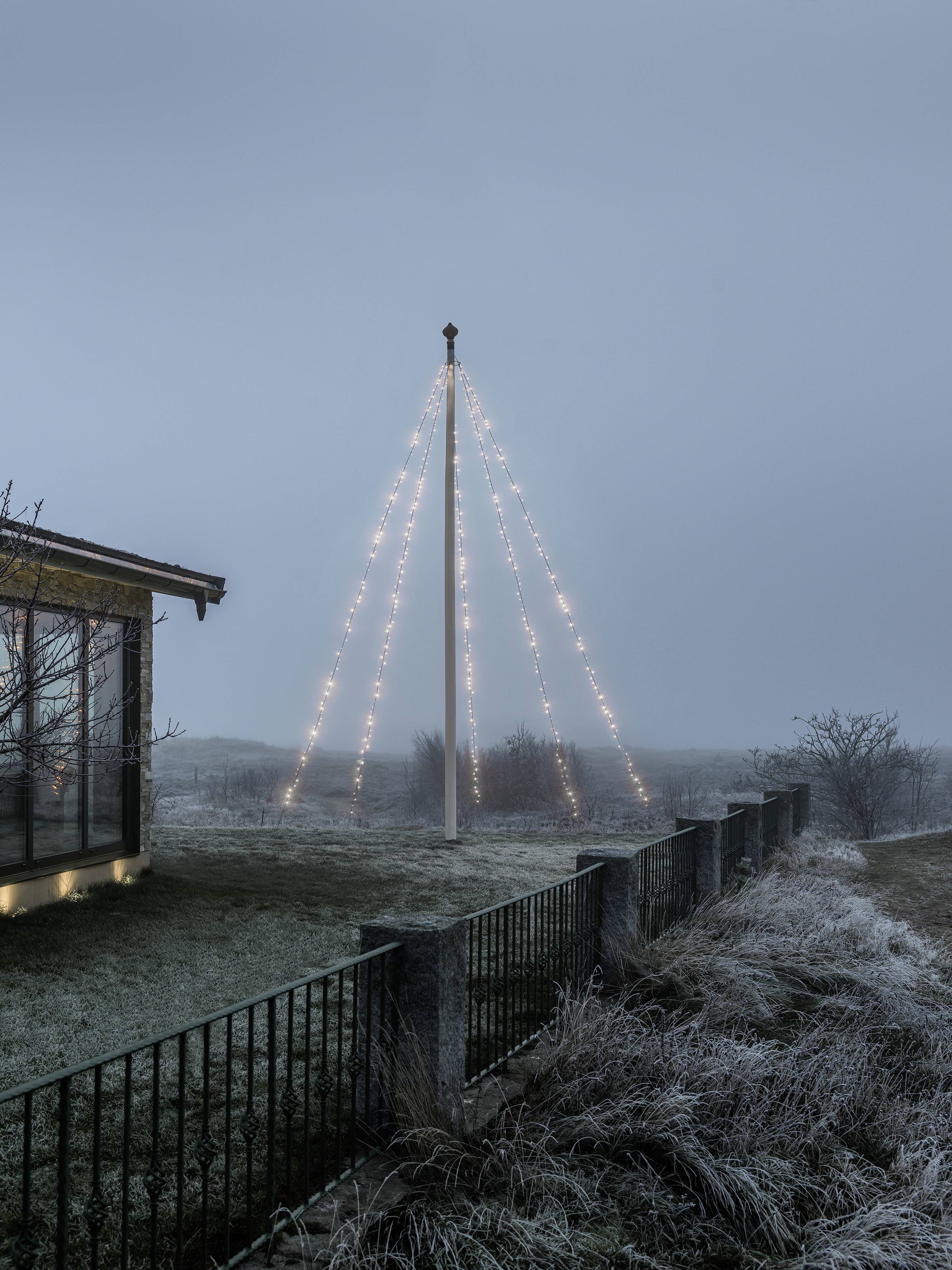 Mast mit Lichterketten in der Dunkelheit auf einer Wiese neben einem Haus. Stimmungsvoll nebelige Umgebung.