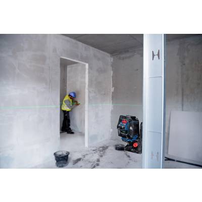 Ein Bauarbeiter im gelben Schutzanzug vermisst eine Wand, während ein Laser-Nivelliergerät zur Messung aufgestellt ist.