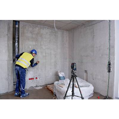 Ein Bauarbeiter mit gelber Weste vermisst eine Betonwand mit einem Lasergerät. Baustelle mit Kabeln und Baumaterialien im Hintergrund.