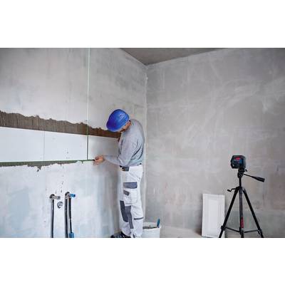 Ein Bauarbeiter mit blauem Helm richtet eine Kachelwand im Rohbau aus. Auf dem Boden steht ein Eimer, daneben ein Stativ mit einem Lasergerät.