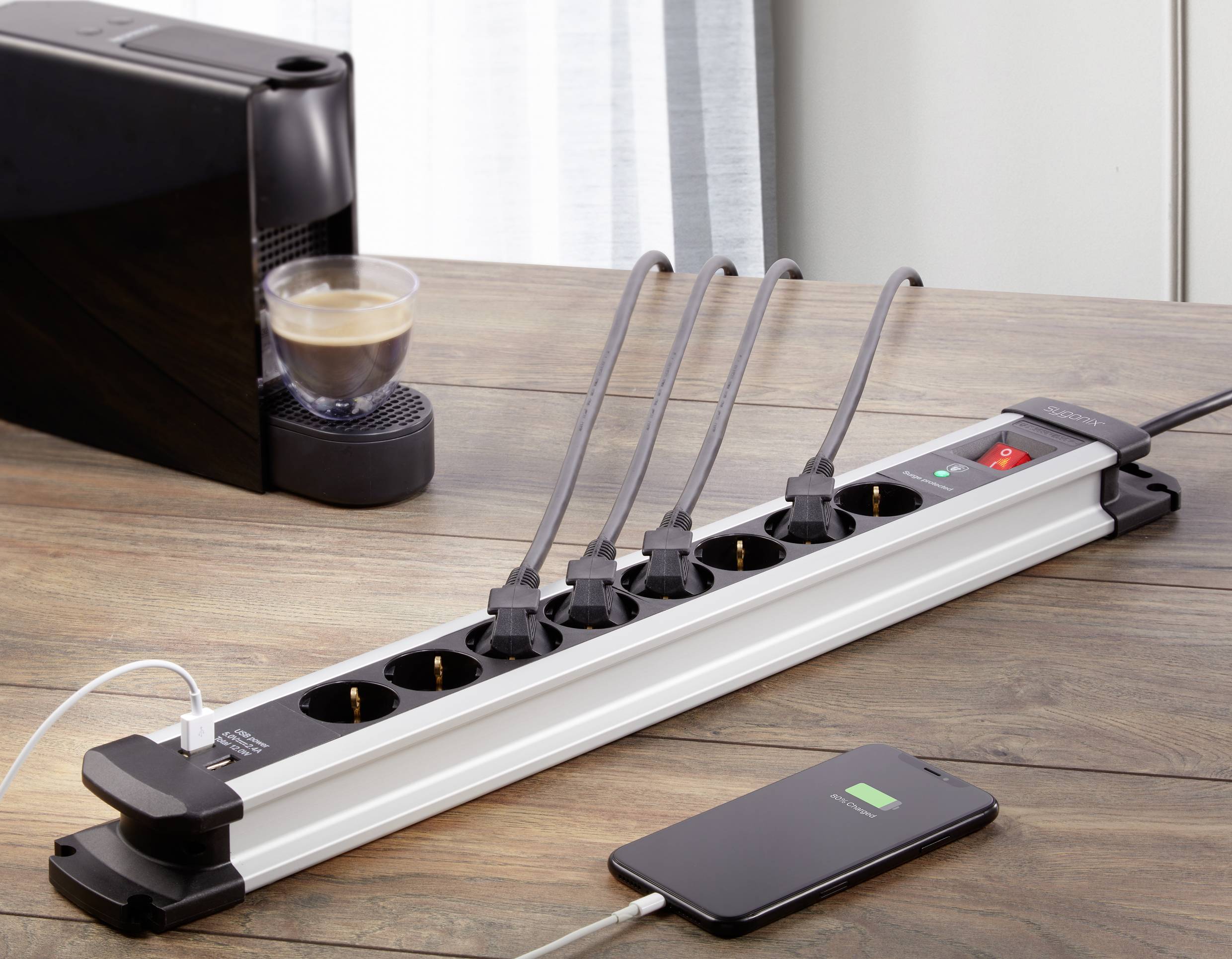 Mehrfachsteckdose mit fünf eingesteckten Kabeln auf Holztisch, daneben Smartphone im Lademodus und Kaffeemaschine mit vollem Glas.