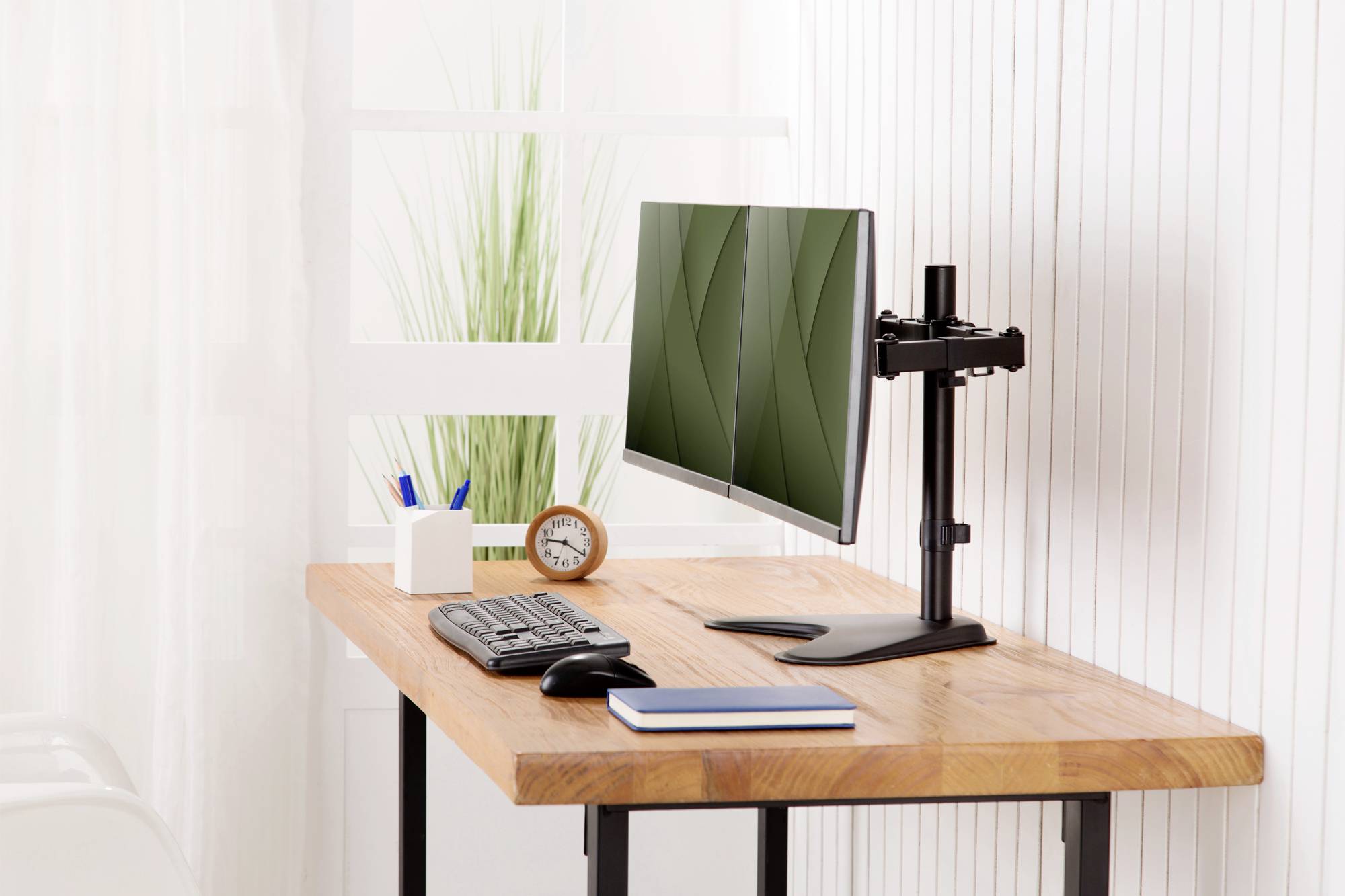 Ein moderner Schreibtisch mit einem Flachbildschirm auf einem Ständer, einer Tastatur, einer Maus, einem Notizbuch und einem Wecker. Pflanzen im Hintergrund.