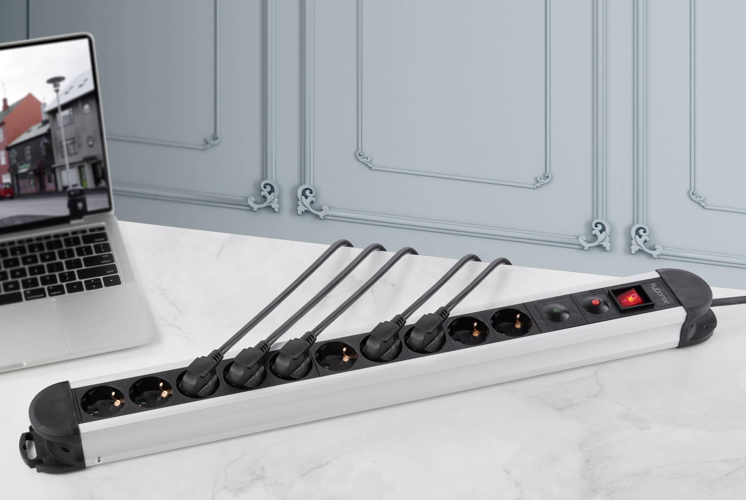 Eine Mehrfachsteckdose mit fünf Steckplätzen und Schalter liegt auf einem Tisch, neben einem geöffneten Laptop, vor blauer Wand.