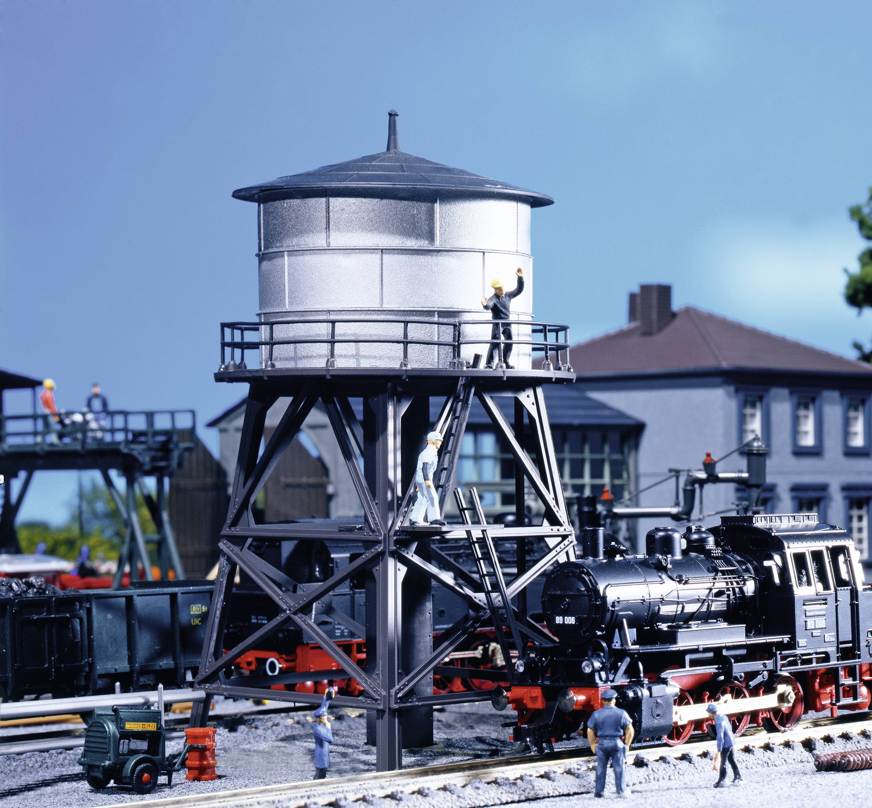 Modellbahnszene mit Dampflokomotive und Wasserturm. Miniaturfiguren arbeiten am Turm. Gebäude und weitere Modelle im Hintergrund.