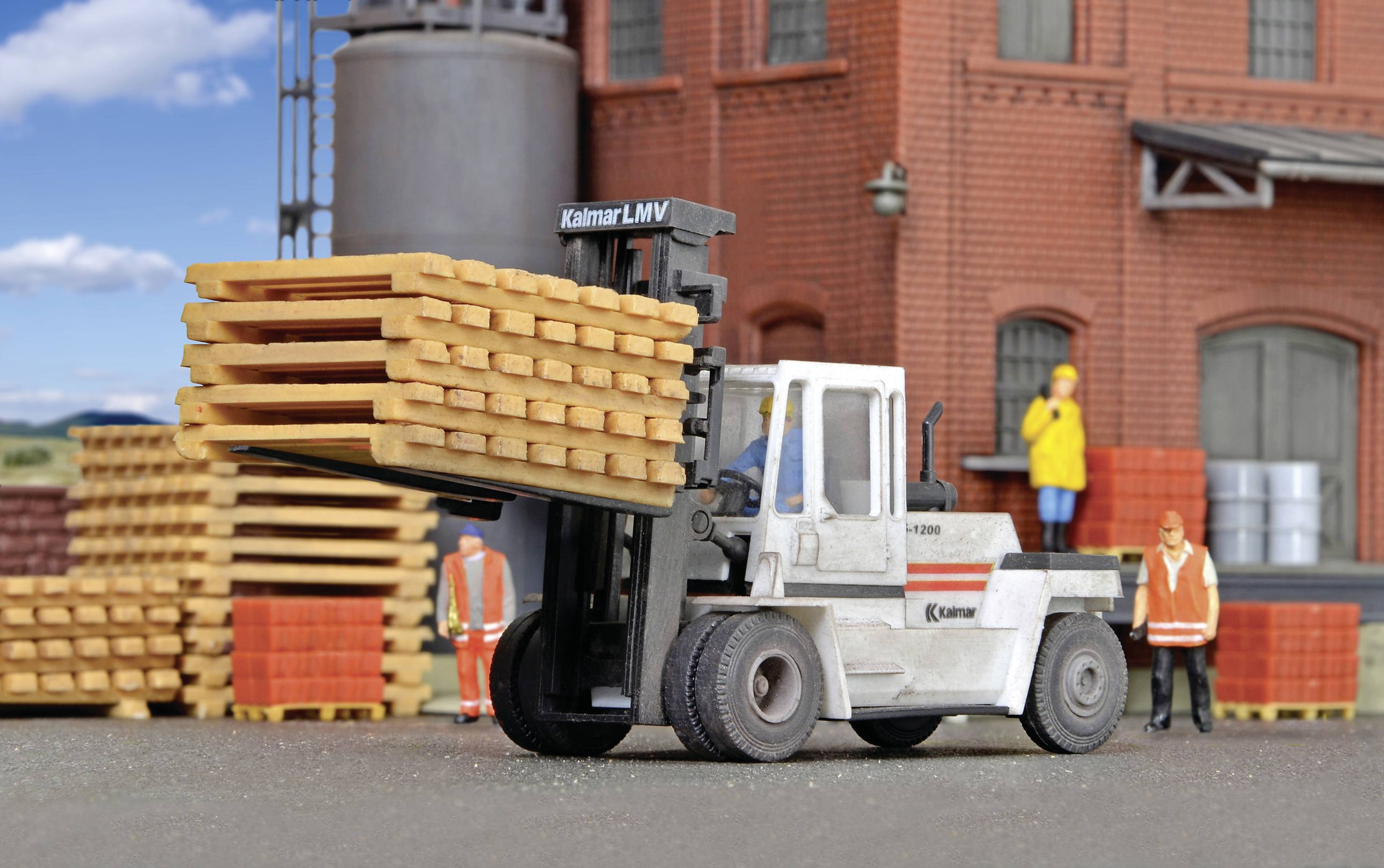 Ein Gabelstapler hebt gestapelte Holzbretter vor einem Backsteingebäude. Zwei Personen in Schutzkleidung stehen in der Nähe.